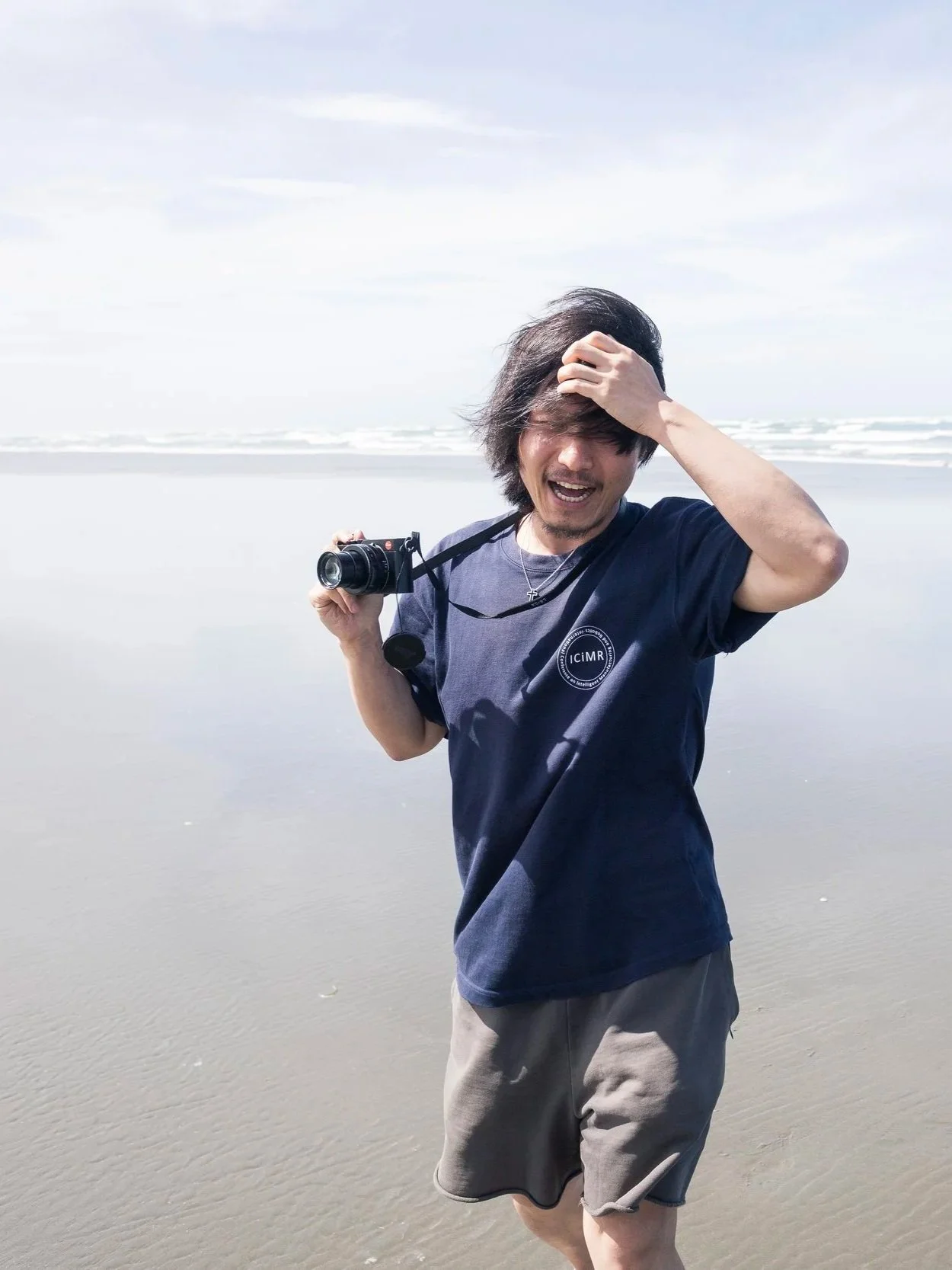 Man standing at the beach holding a camera, with windblown hair, smiling and touching his forehead, wearing a dark t-shirt and gray shorts.