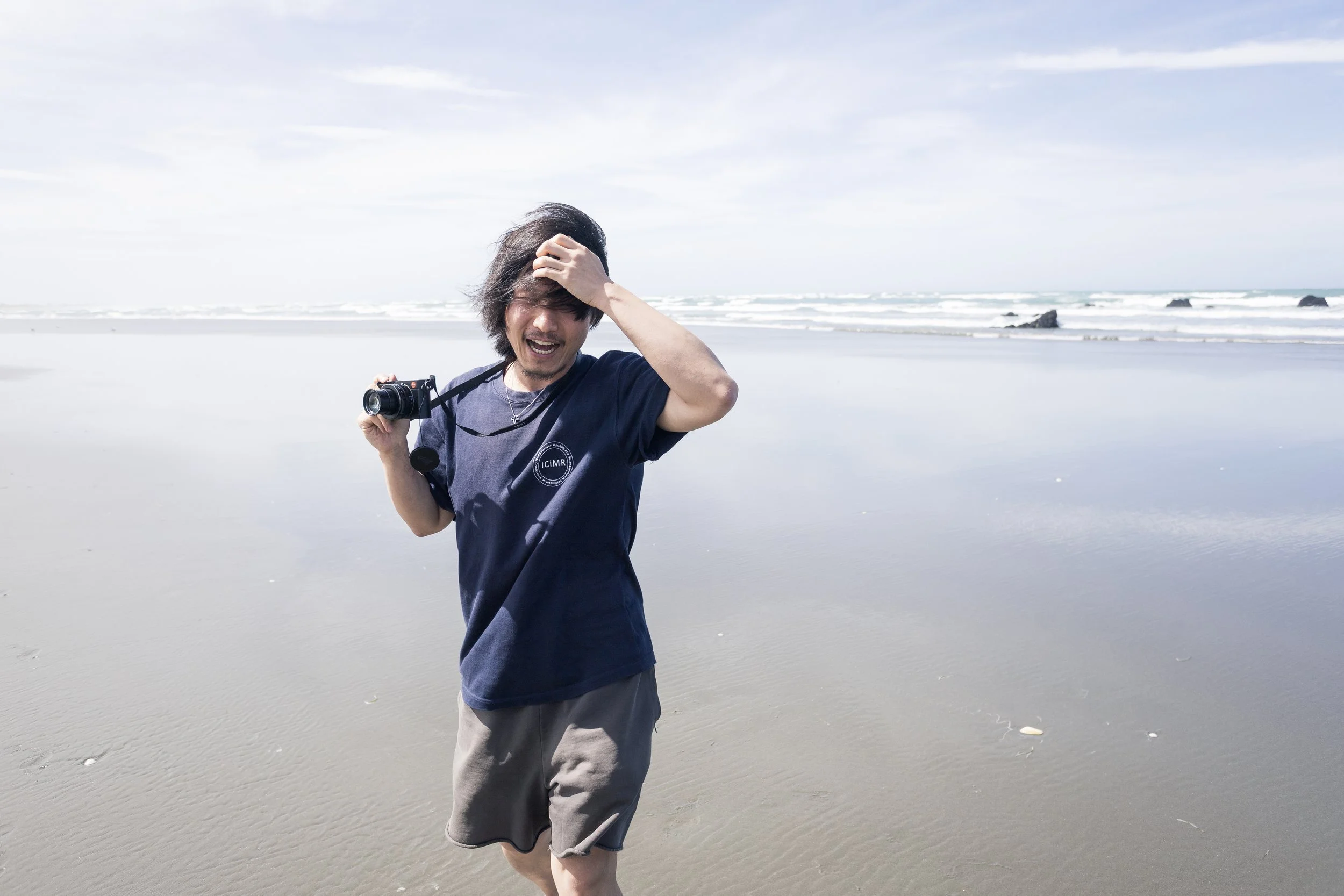 A man on the beach holding a camera and playing with his hair, walking near the water.