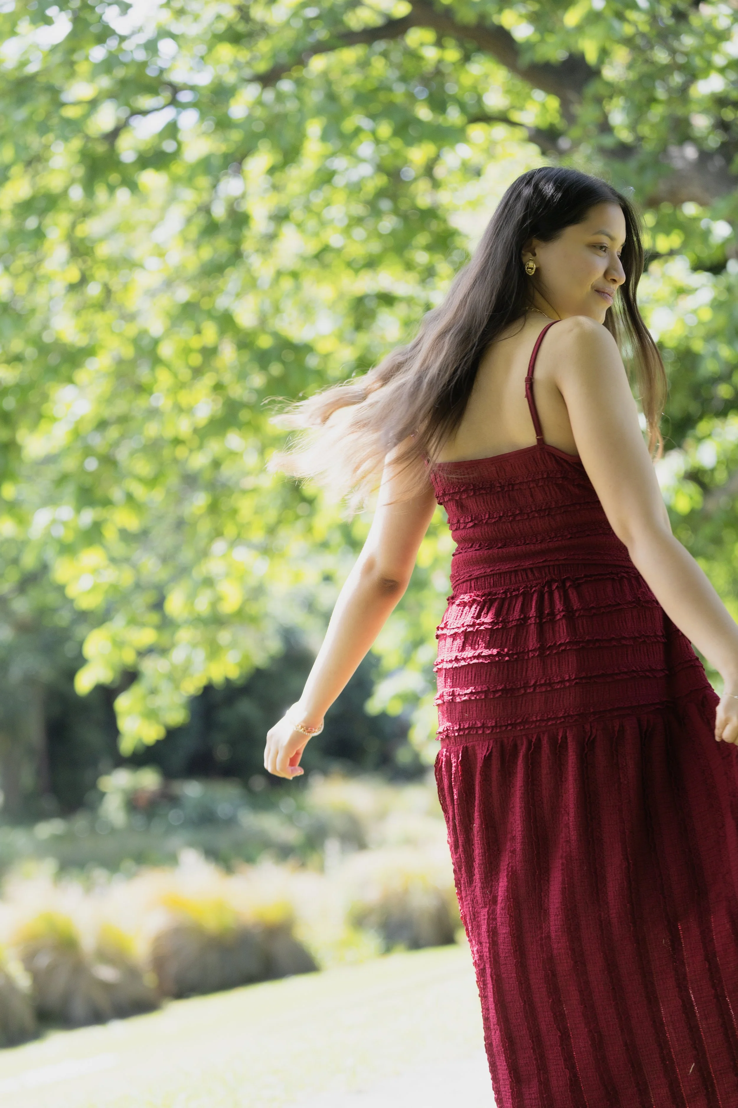 A woman in a red dress standing outdoors among green trees with sunlight filtering through the leaves.