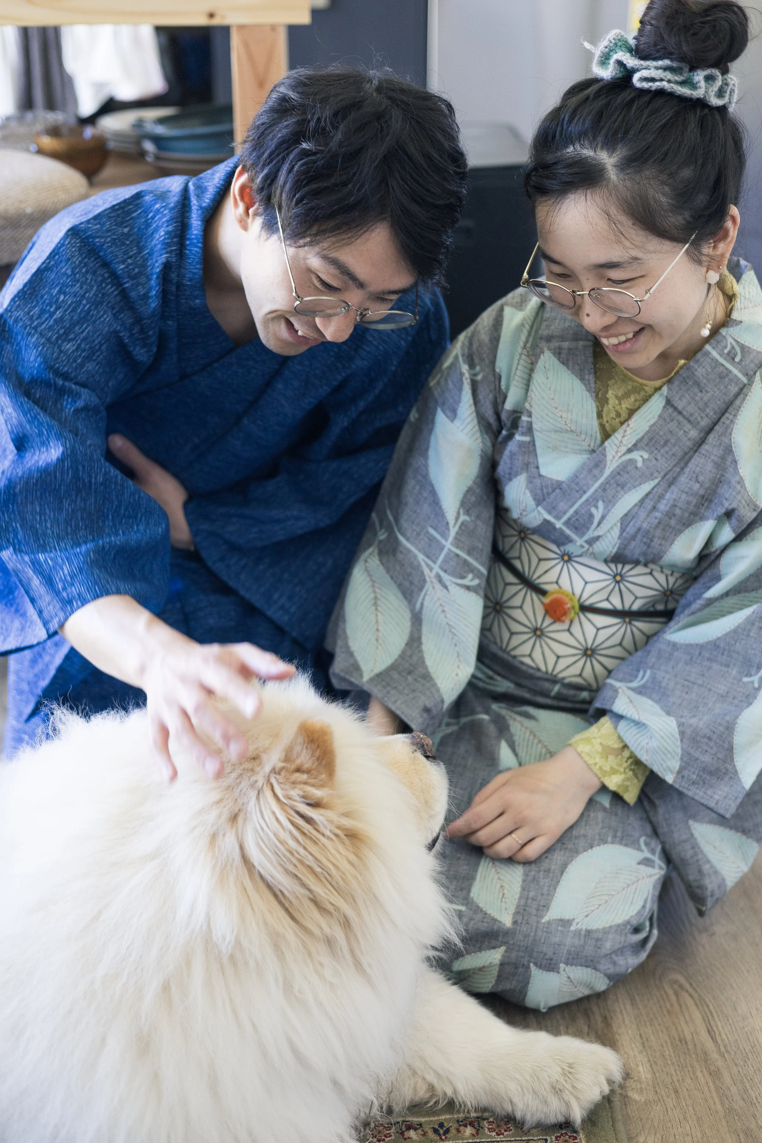 Two people in traditional Japanese attire pet a large, fluffy dog lying on the wooden floor indoors.