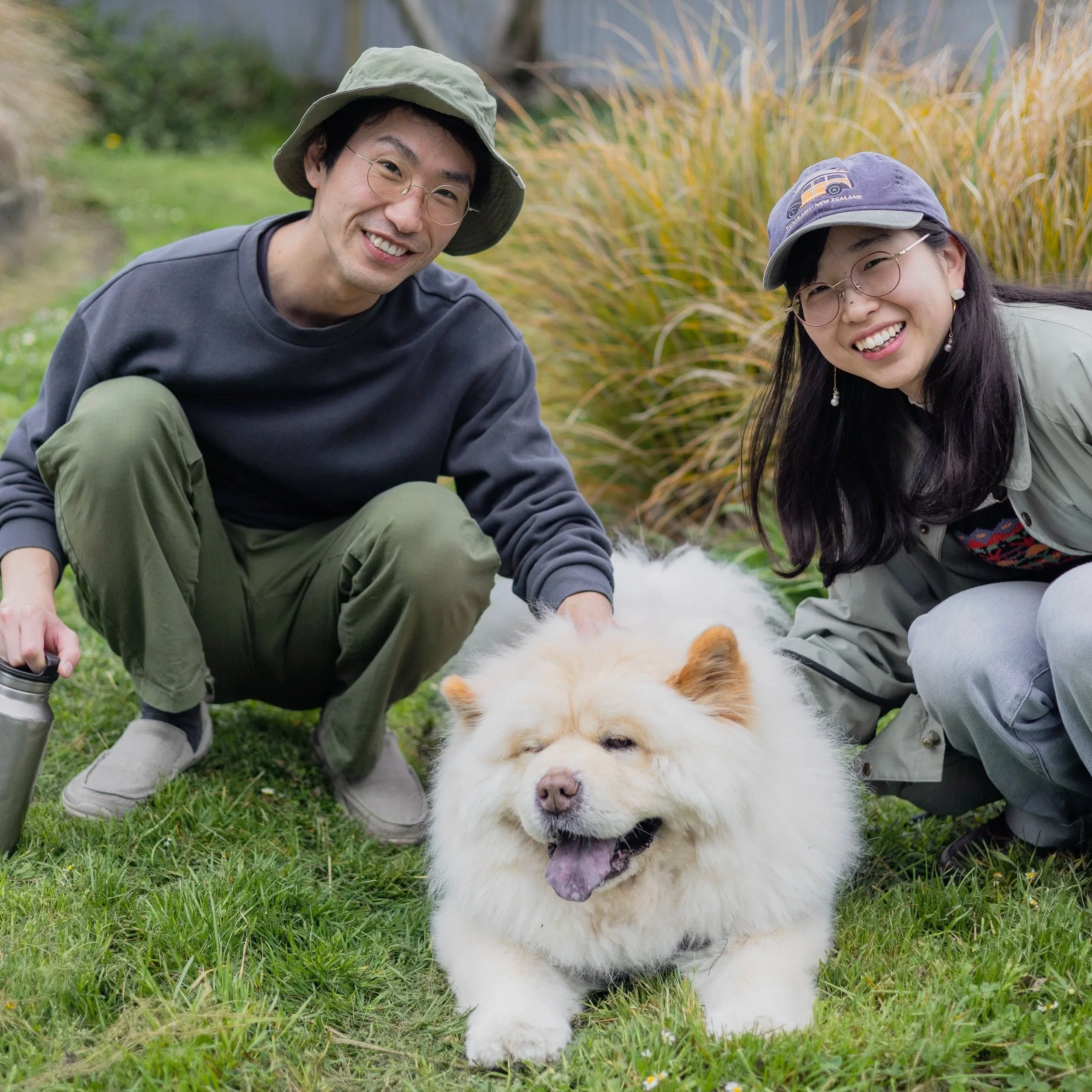 A smiling man and woman outdoors squatting next to a fluffy white dog with a thick coat, in a grassy area with tall grass in the background.