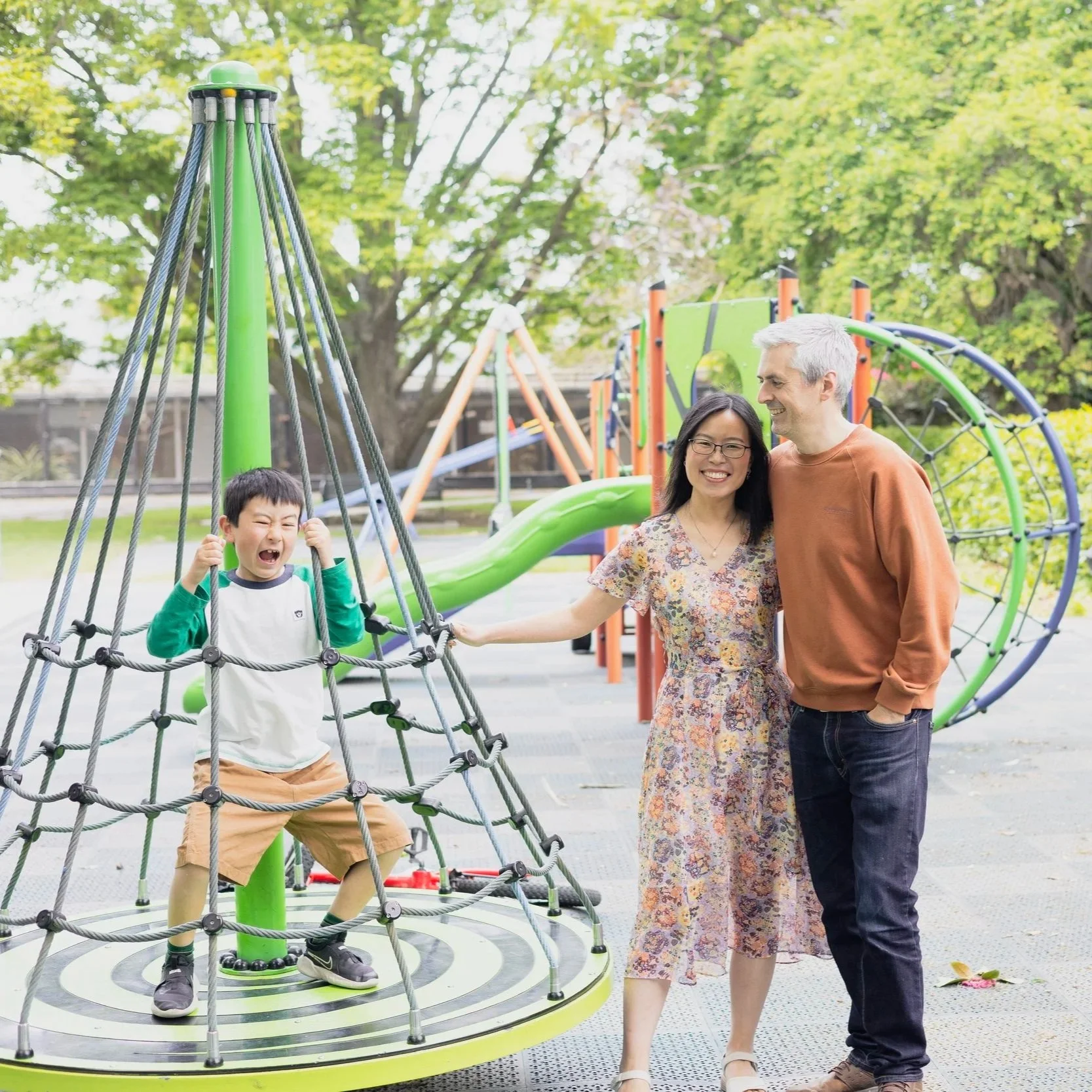 A boy laughing on a playground climbing structure, three adults nearby, two women and one man, smiling and enjoying a moment together, with trees and playground equipment in the background.
