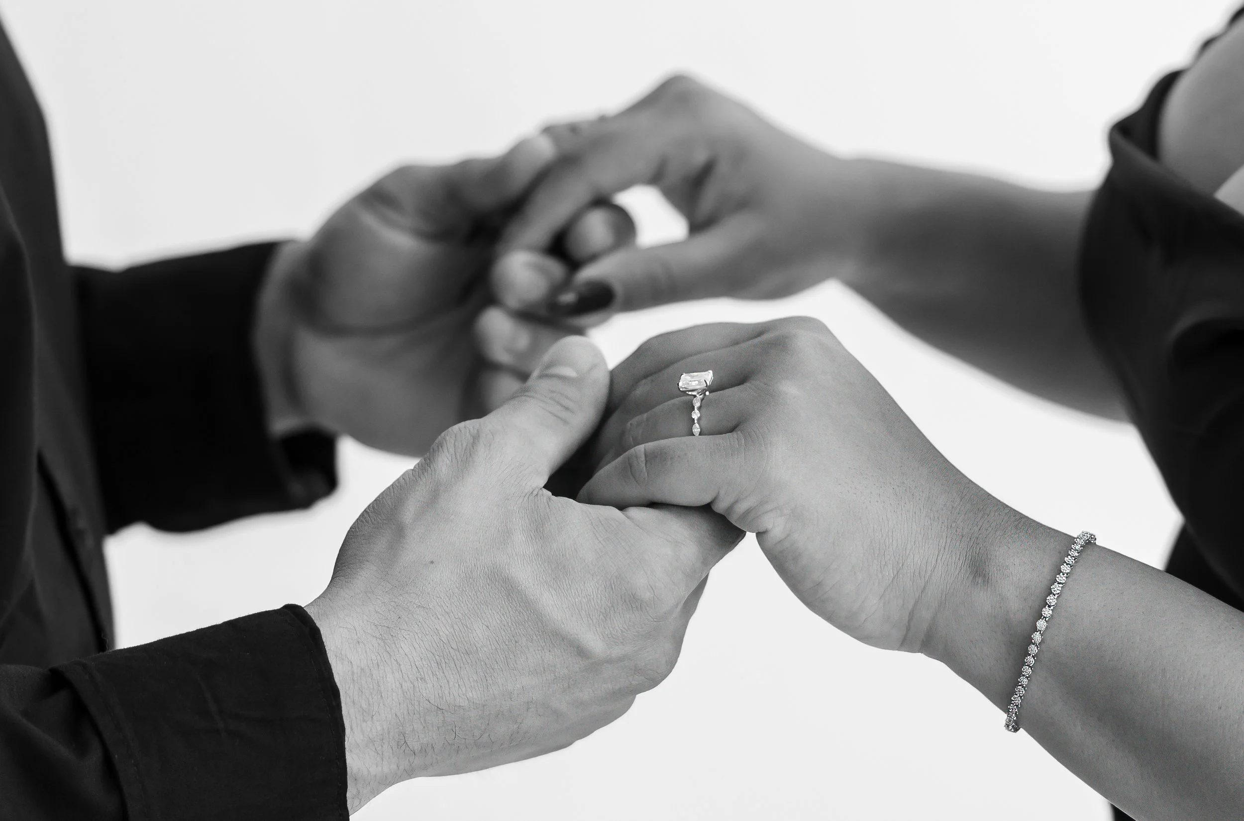 Close-up of hands exchanging rings, showing a diamond engagement ring on one hand, with a person wearing a bracelet. The photo is in black and white.