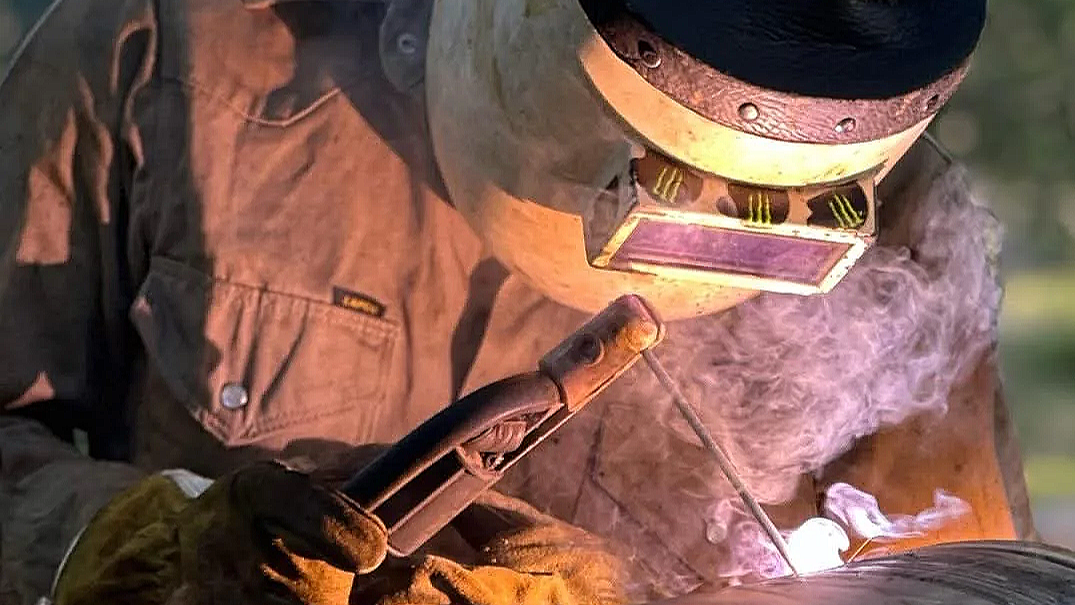 A person wearing protective welding gear, including a helmet with a pink filter, is welding metal outdoors. Sparks and smoke are visible.