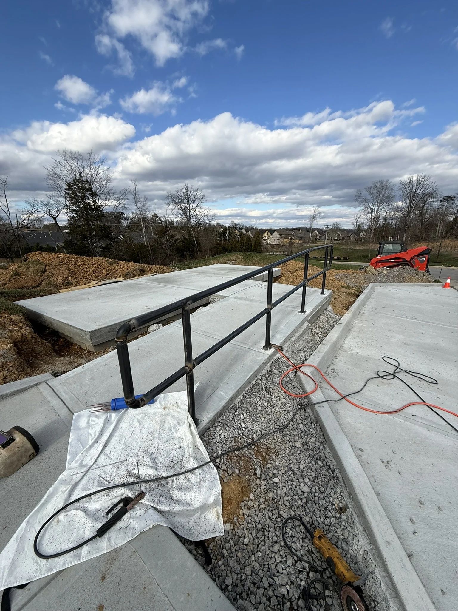 Construction site showing a freshly poured concrete sidewalk with a black metal handrail, construction tools, and equipment, under a cloudy sky.
