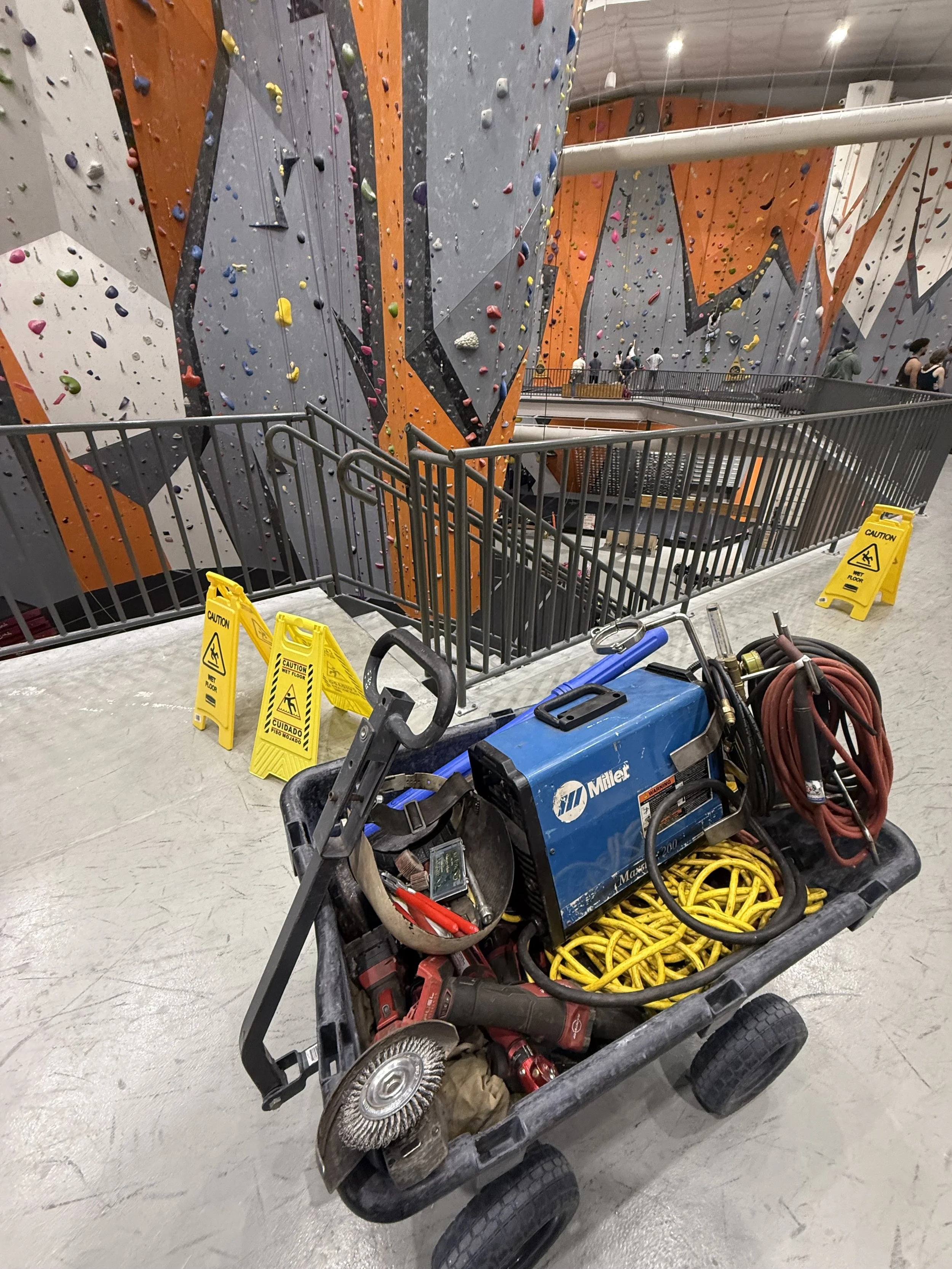 Indoor rock climbing gym with artificial climbing walls in orange, gray, and white colors, some people climbing and others observing. In the foreground, a cart with various tools and equipment such as a blue Miller welder, extension cords, a grinder, and safety signs indicating wet floor.