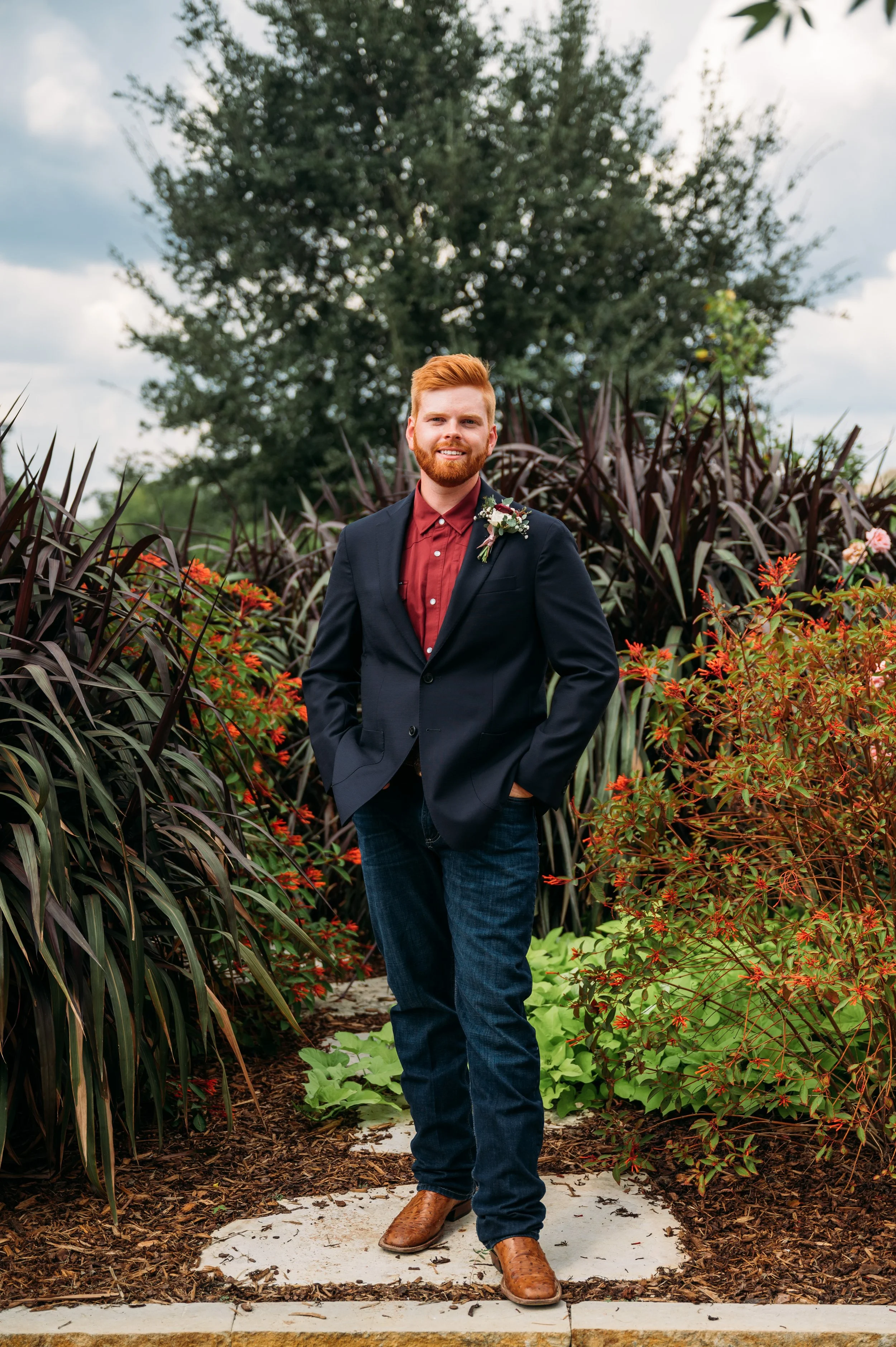 A man with red hair and beard standing outdoors on a small stone path, wearing a navy blazer, burgundy shirt, dark jeans, and brown boots, with a boutonniere on his blazer, surrounded by colorful bushes and trees.