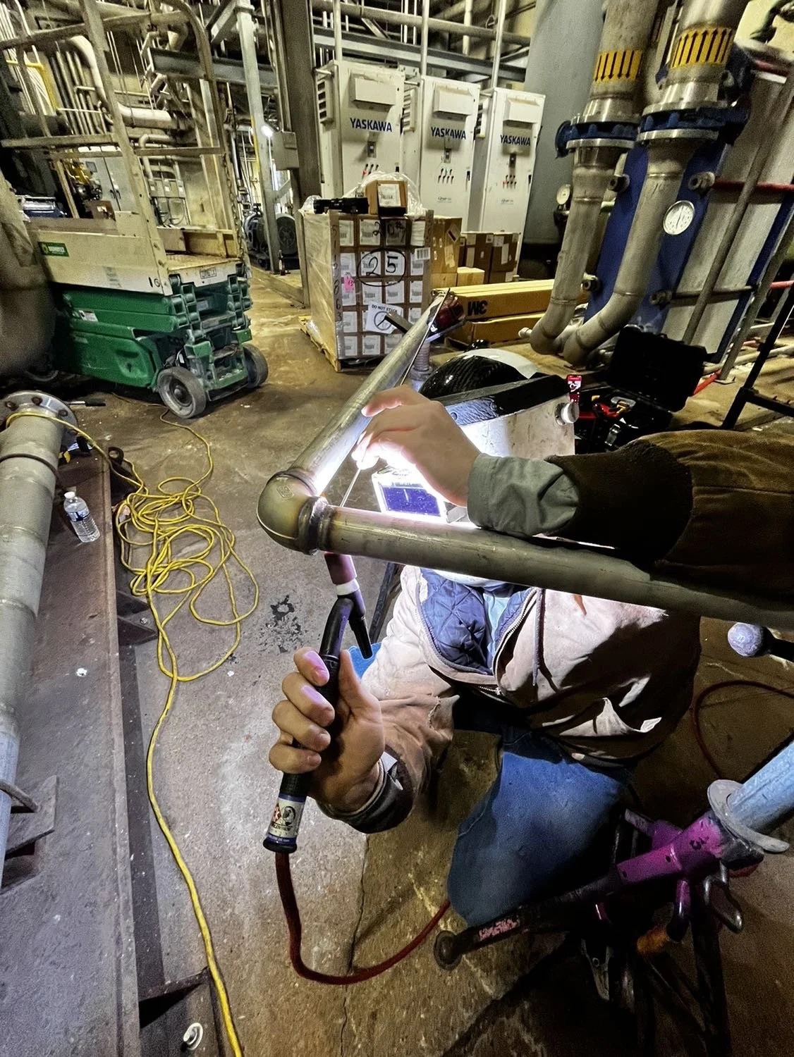 A worker welding a metal pipe in an industrial facility. The worker is kneeling on the ground, holding a welding torch with bright sparks visible. The background includes various pipes, control boxes, and boxes of supplies, with yellow extension cords on the floor.