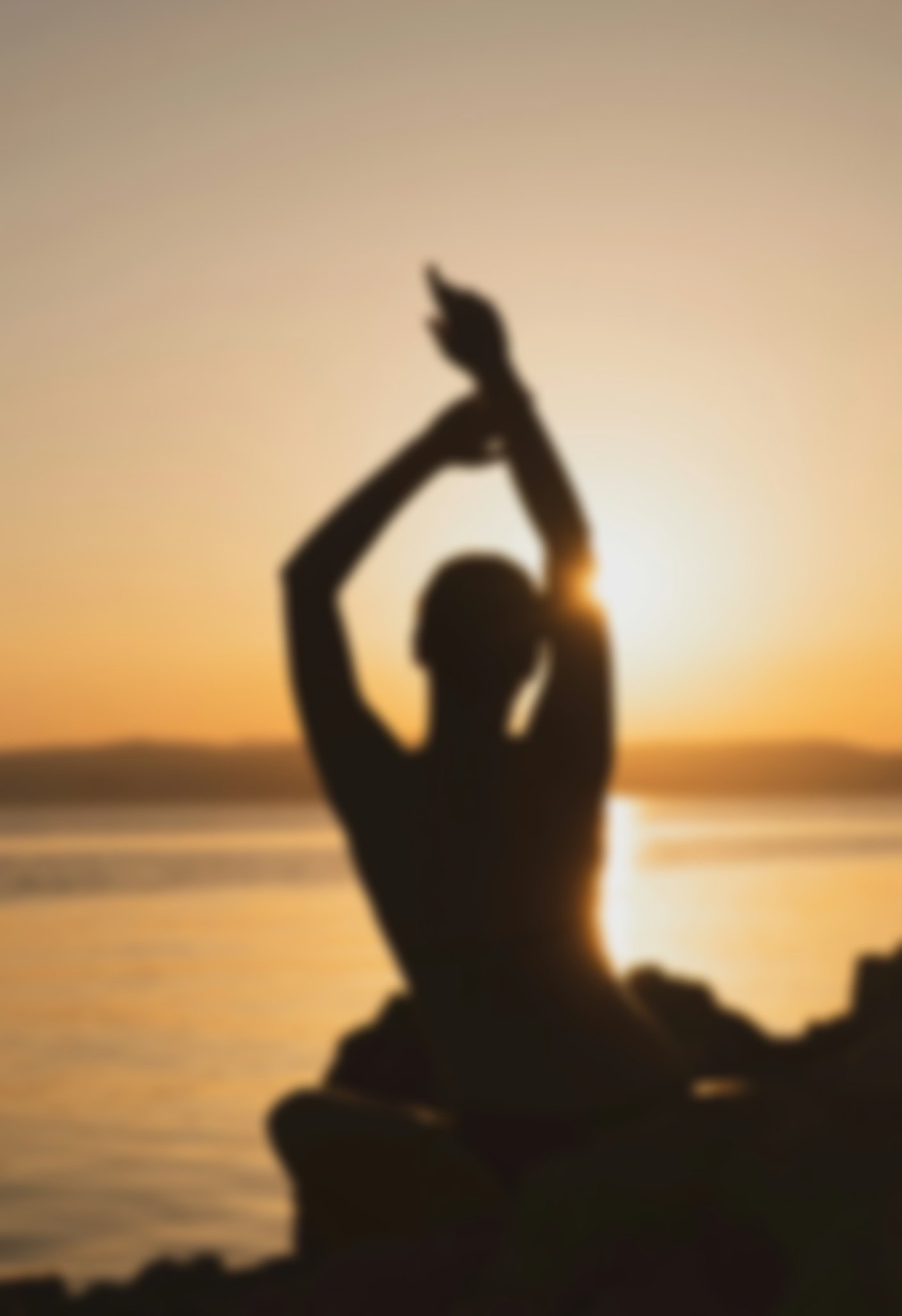 Silhouette of a woman with arms raised, sitting on rocks at sunset over water.