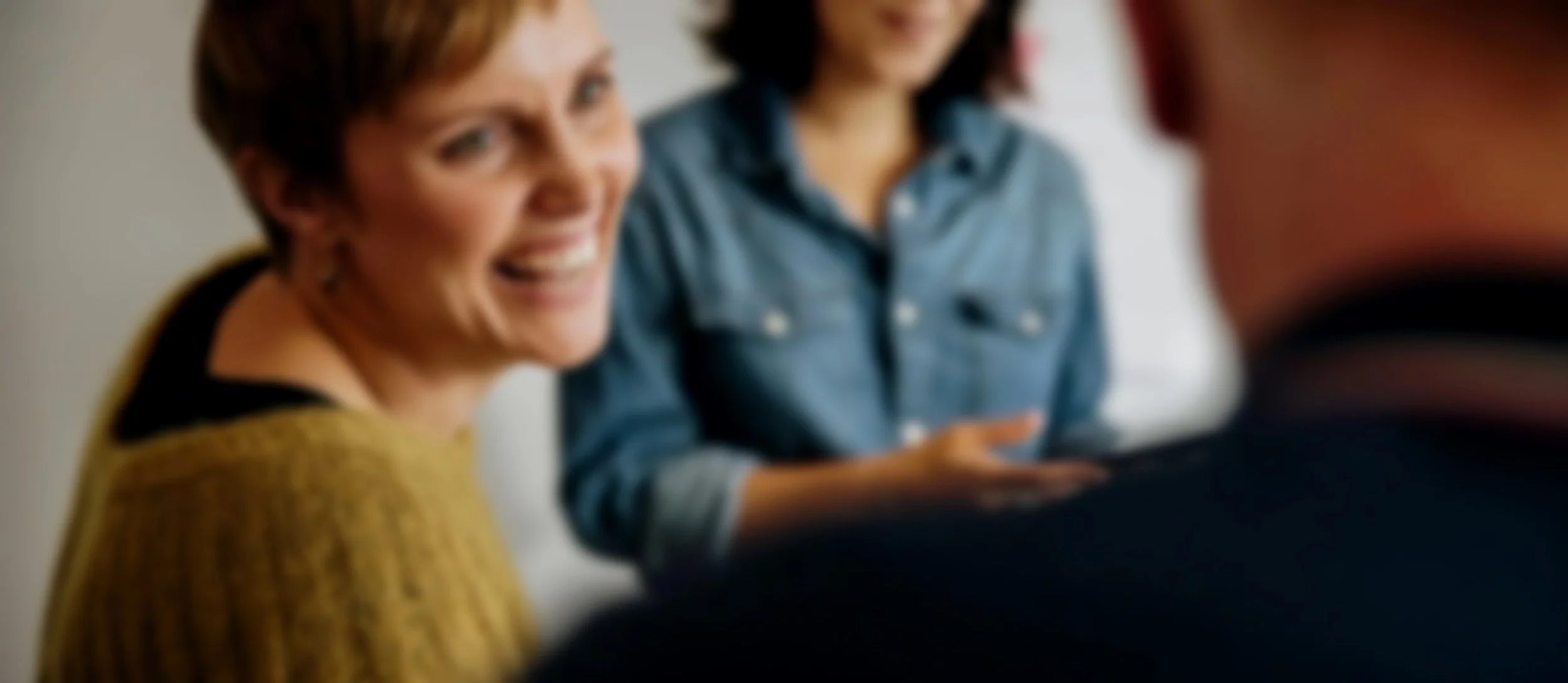 A woman with short hair smiling at the camera, with two other women blurred in the background, engaged in conversation.