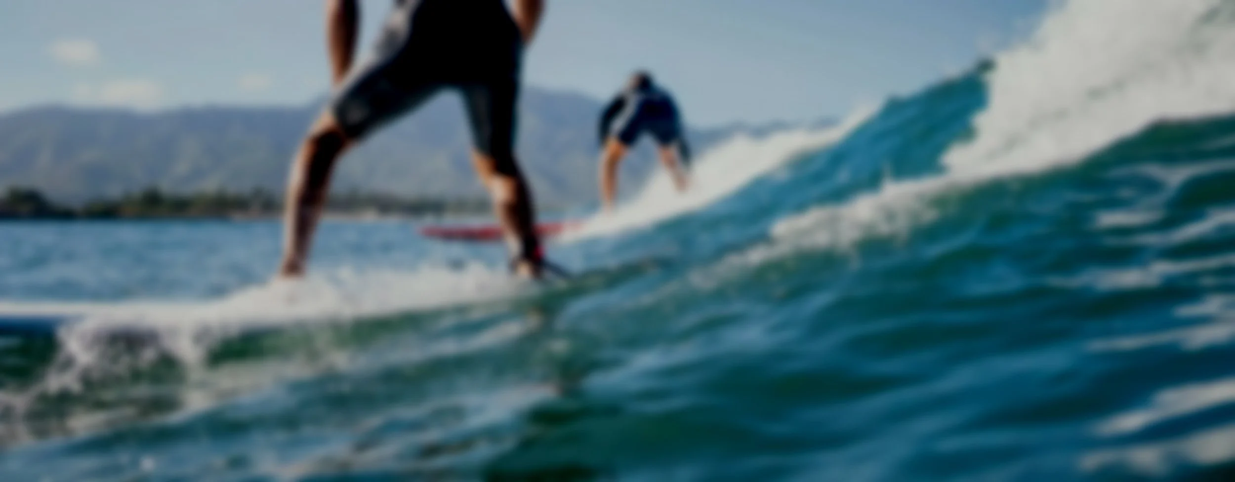 Two people surfing on ocean waves near a mountainous coastline under a partly cloudy sky.