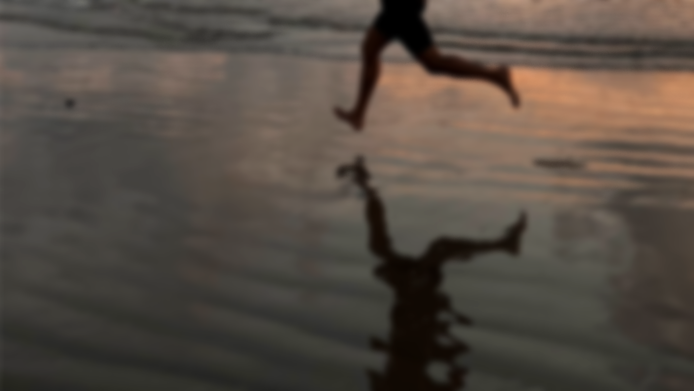 A person playing or jumping on the wet sand at the beach during sunset, with their reflection visible in the shallow water.
