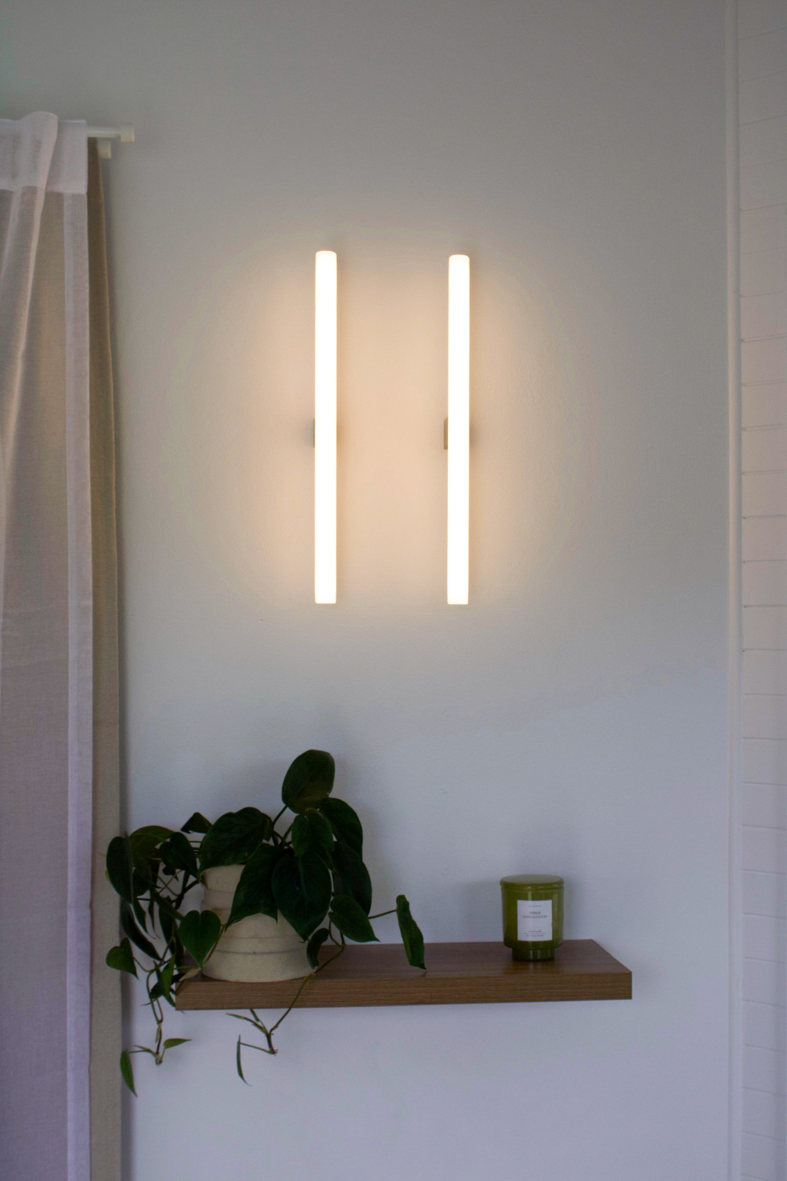 Wall-mounted modern lamp with two vertical light bars, a green candle, and a potted plant on a wooden shelf.