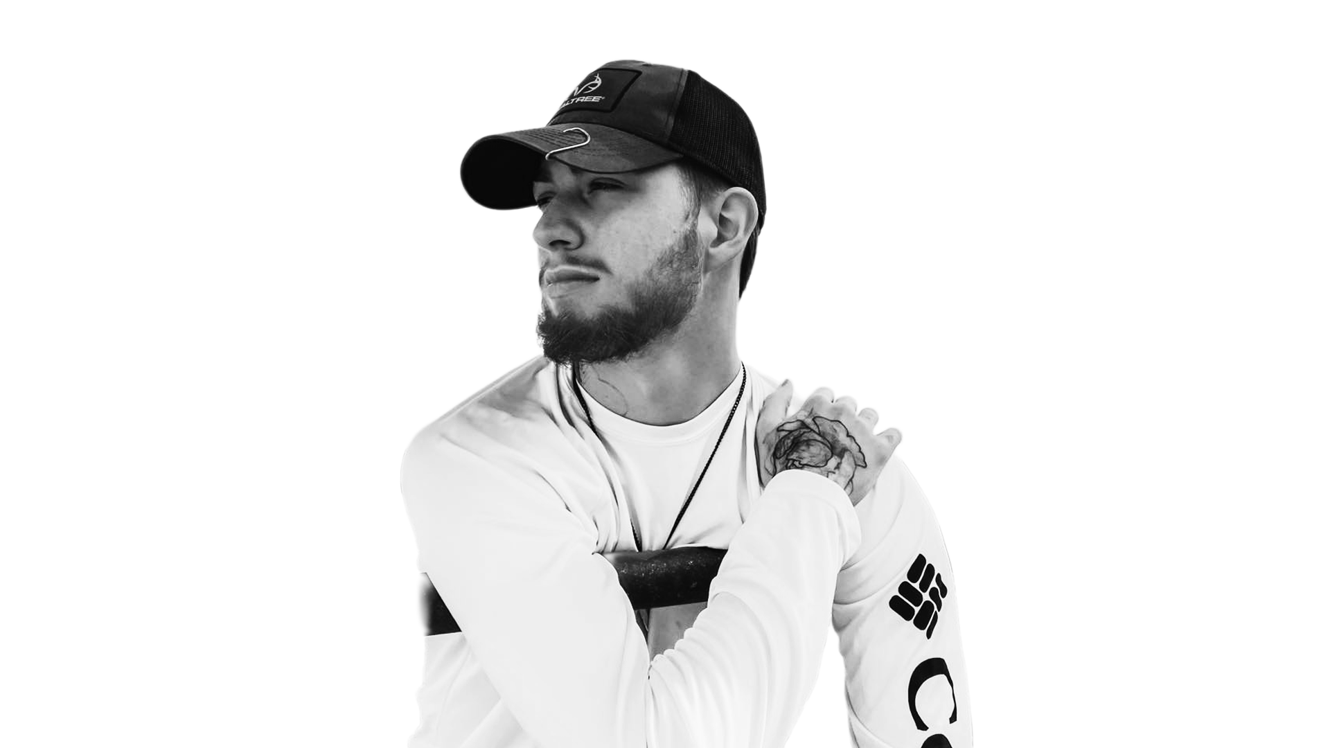 Black and white photo of a young man with a beard, wearing a baseball cap, looking to the side, with his right hand resting on his shoulder. He has tattoos on his left arm and is wearing a long-sleeve white shirt with logos.