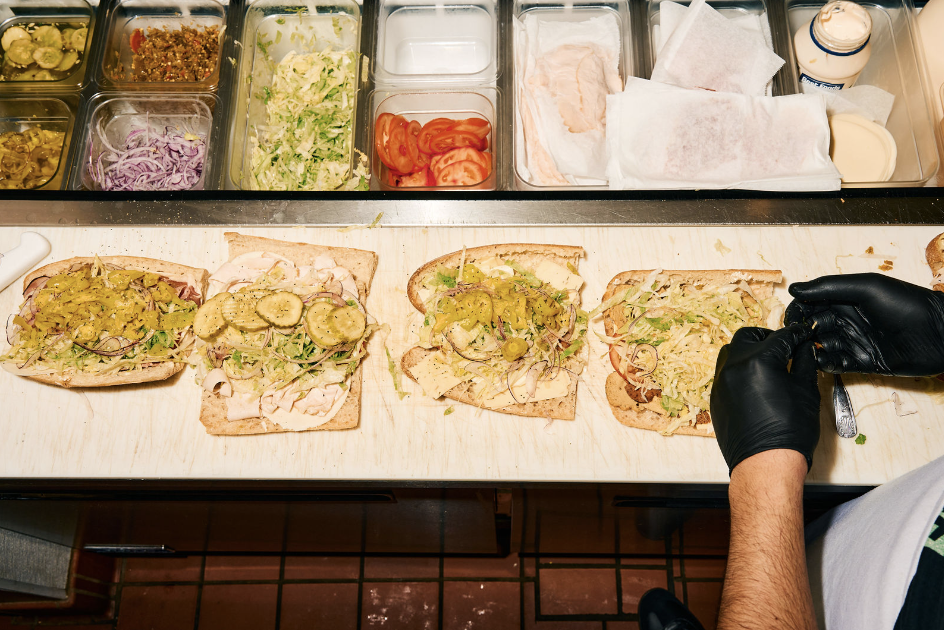 A worker prepares hoagies with pickles, lettuce, tomatoes, and other ingredients on a wooden counter, with a variety of toppings in containers above.