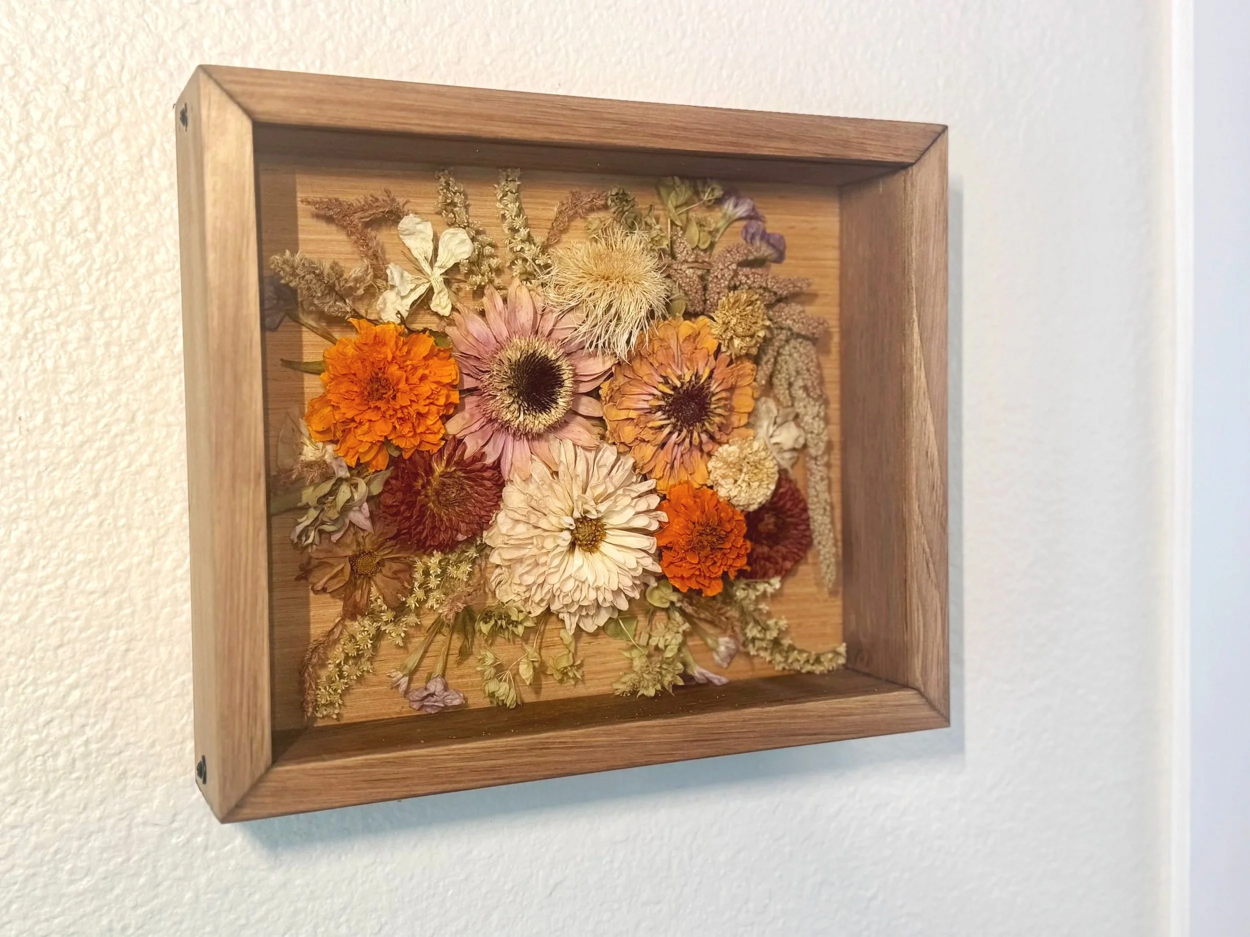 A shadow box wall art filled with dried multicolored flowers and greenery, framed in wood, hanging on a white wall.