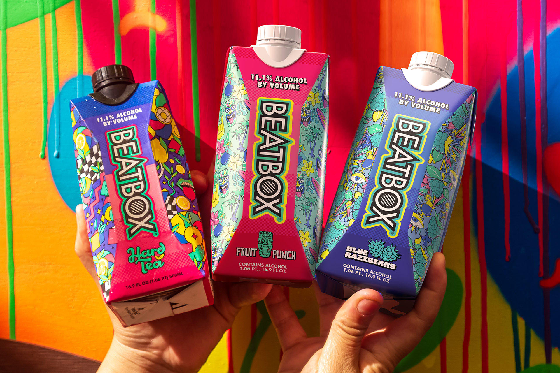 Three hands holding colorful boxes of BEARBOX alcoholic beverages against a vibrant, painted background with drips of paint.