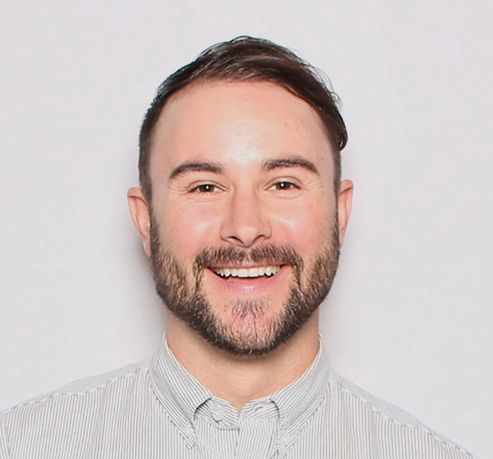 Portrait of a smiling man with short brown hair, beard, and light-colored striped shirt against a plain background.