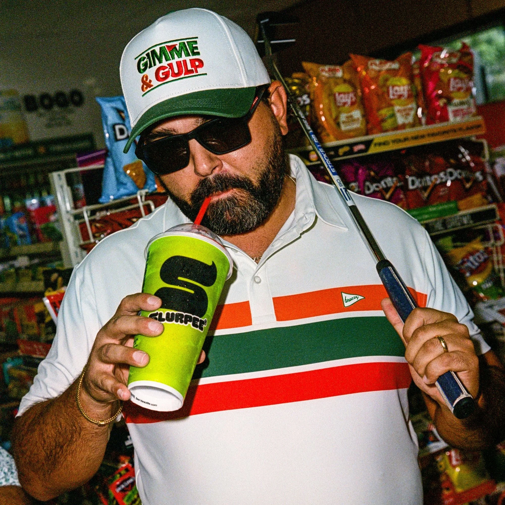 A man with a beard dressed in a white shirt with red and green stripes, wearing sunglasses and a cap that reads 'Gimme & Gulp', is sipping from a large green cup with a straw at a convenience store. He is holding a golf club in his left hand.