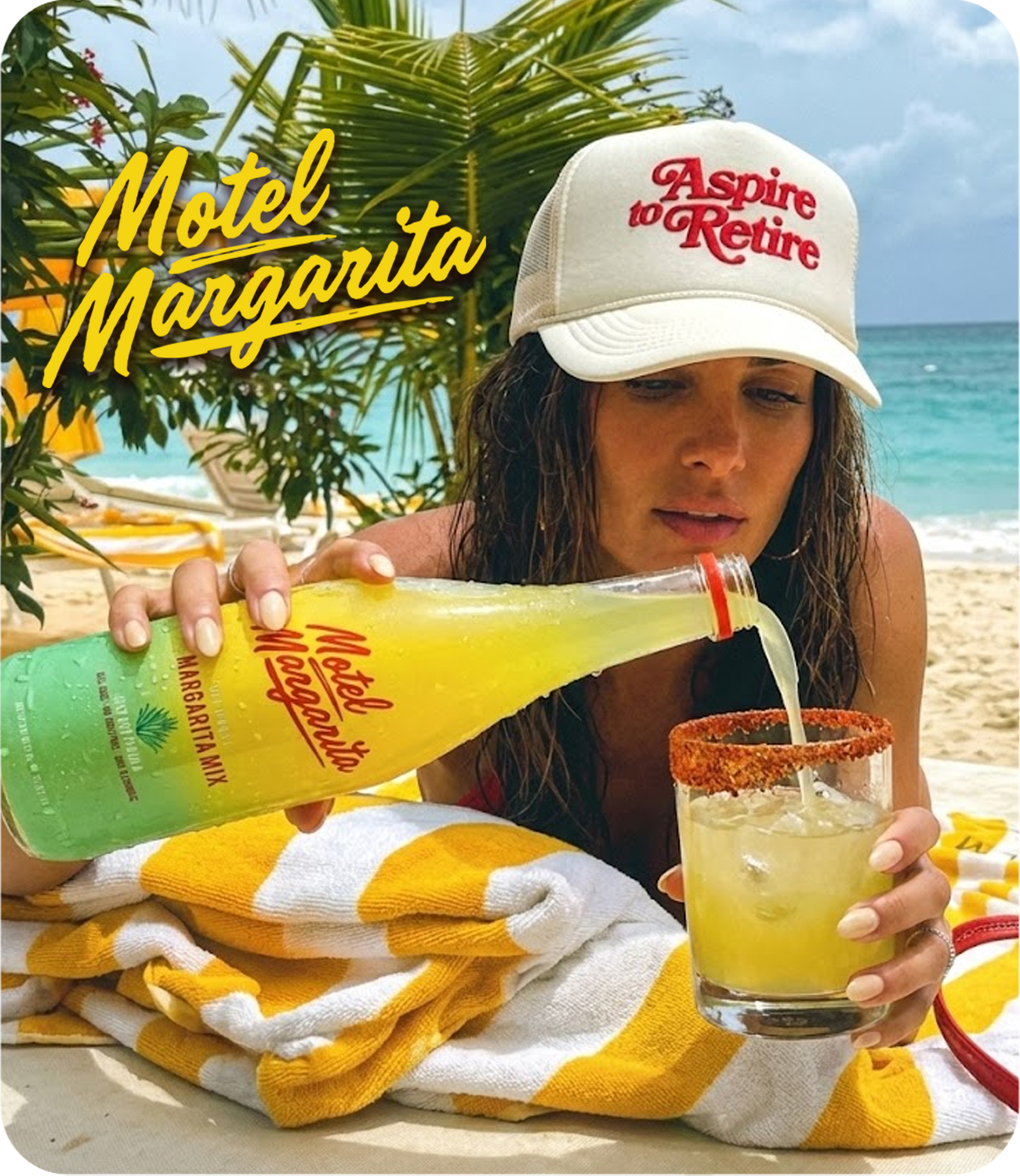 A woman with wet hair wearing a white cap with red text, pouring a yellow drink from a green bottle into a glass with a salted rim on a beach. There are palm leaves and the ocean in the background, with yellow towels or clothing nearby.