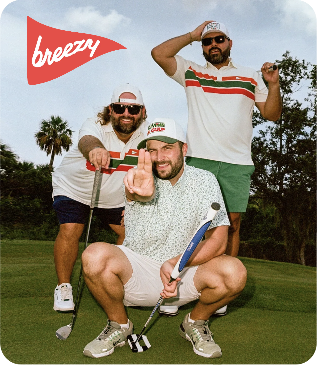 Three men playing golf on a course, wearing casual golf attire, with one squatting in front holding a golf club and making a peace sign, and two standing behind, one with hand on his head and the other with hand on his shoulder, all smiling. A red pennant with 'breezy' written in white is in the top left corner.