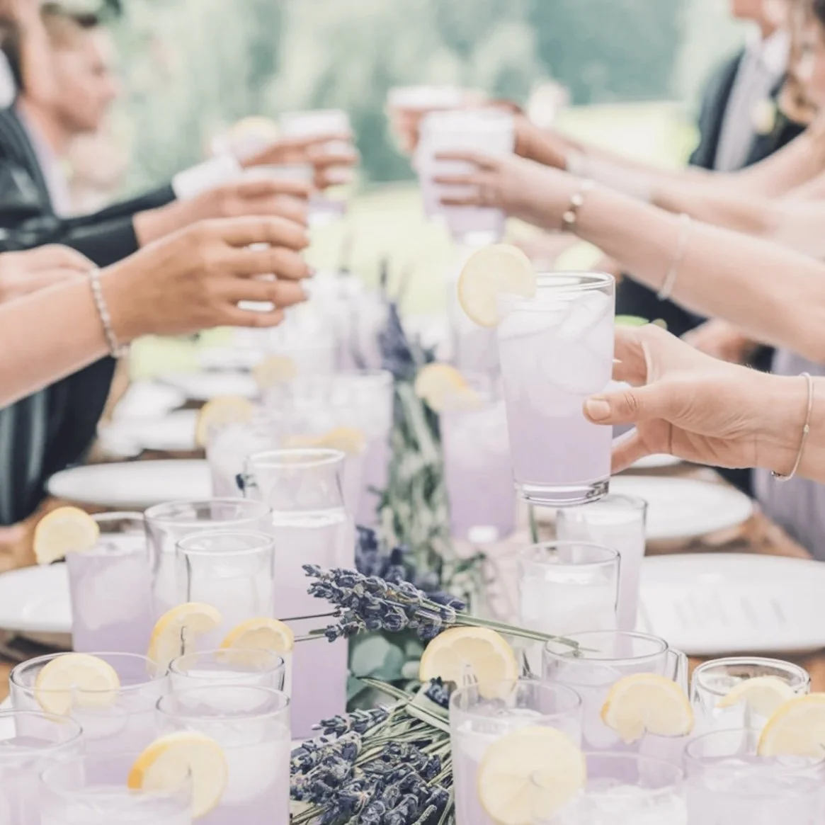 A group of people at an outdoor celebration raising glasses of lemonade with lemon slices, with a lavender centerpiece on the table.