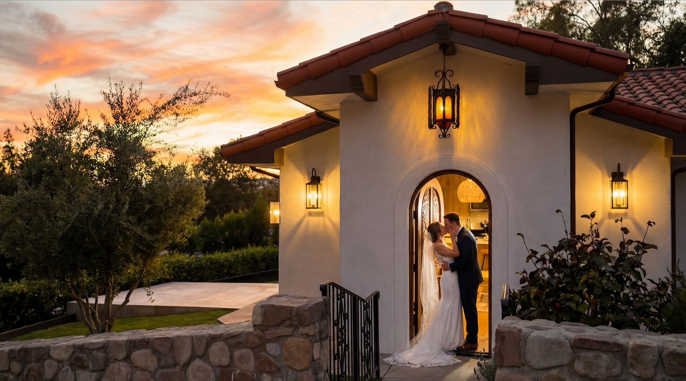 A couple dressed in wedding attire sharing a kiss at the entrance of a small, charming house at sunset, with warm lighting and a colorful sky.