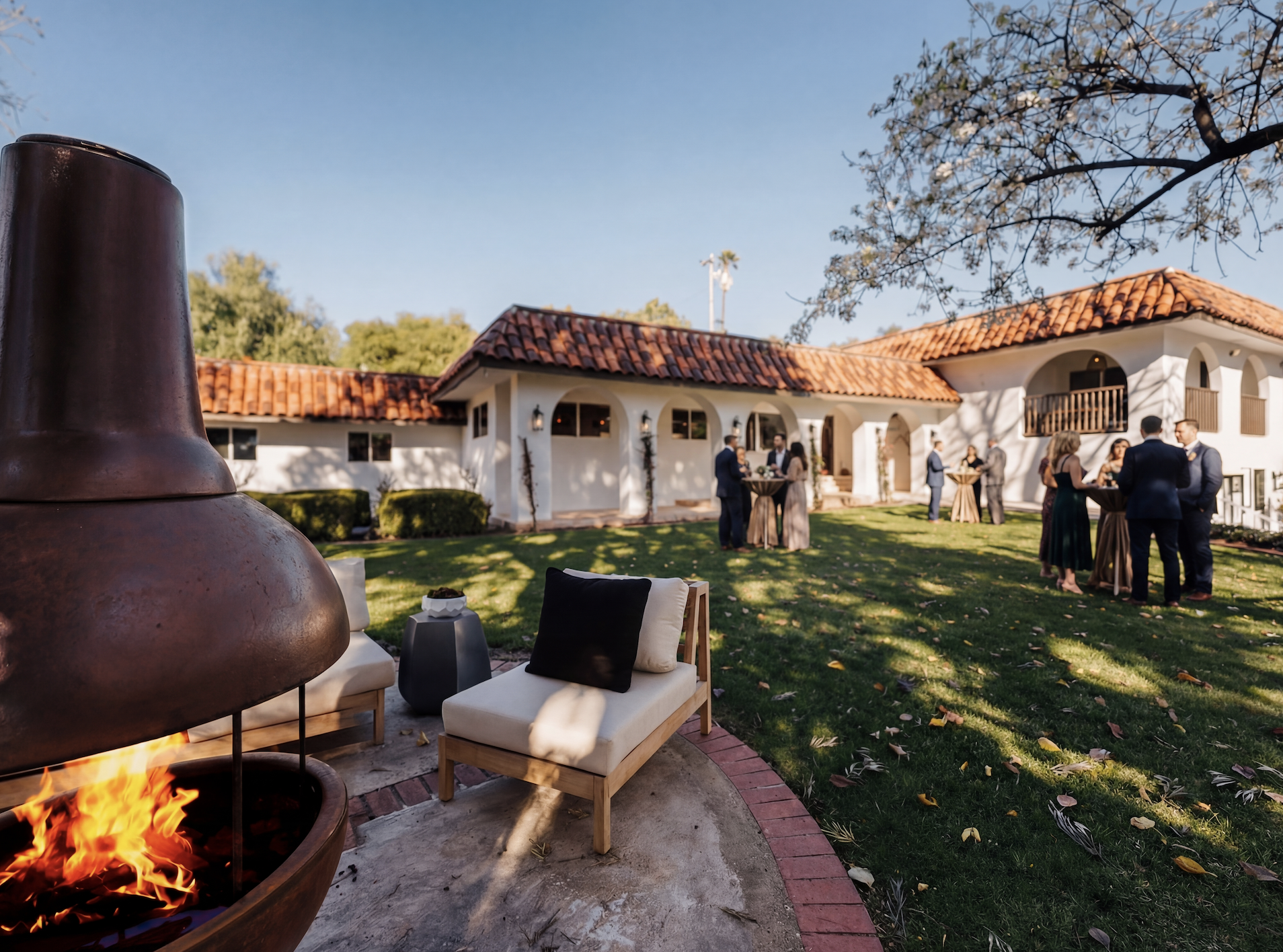 Outdoor social gathering with people standing around tall tables in the backyard of a house with Spanish-style architecture, red tile roof, and white stucco walls. There is a fire pit with a chimenea and seating area with a chair and pillows in the foreground. The scene is set on a sunny day with shadows cast on the grass and trees.