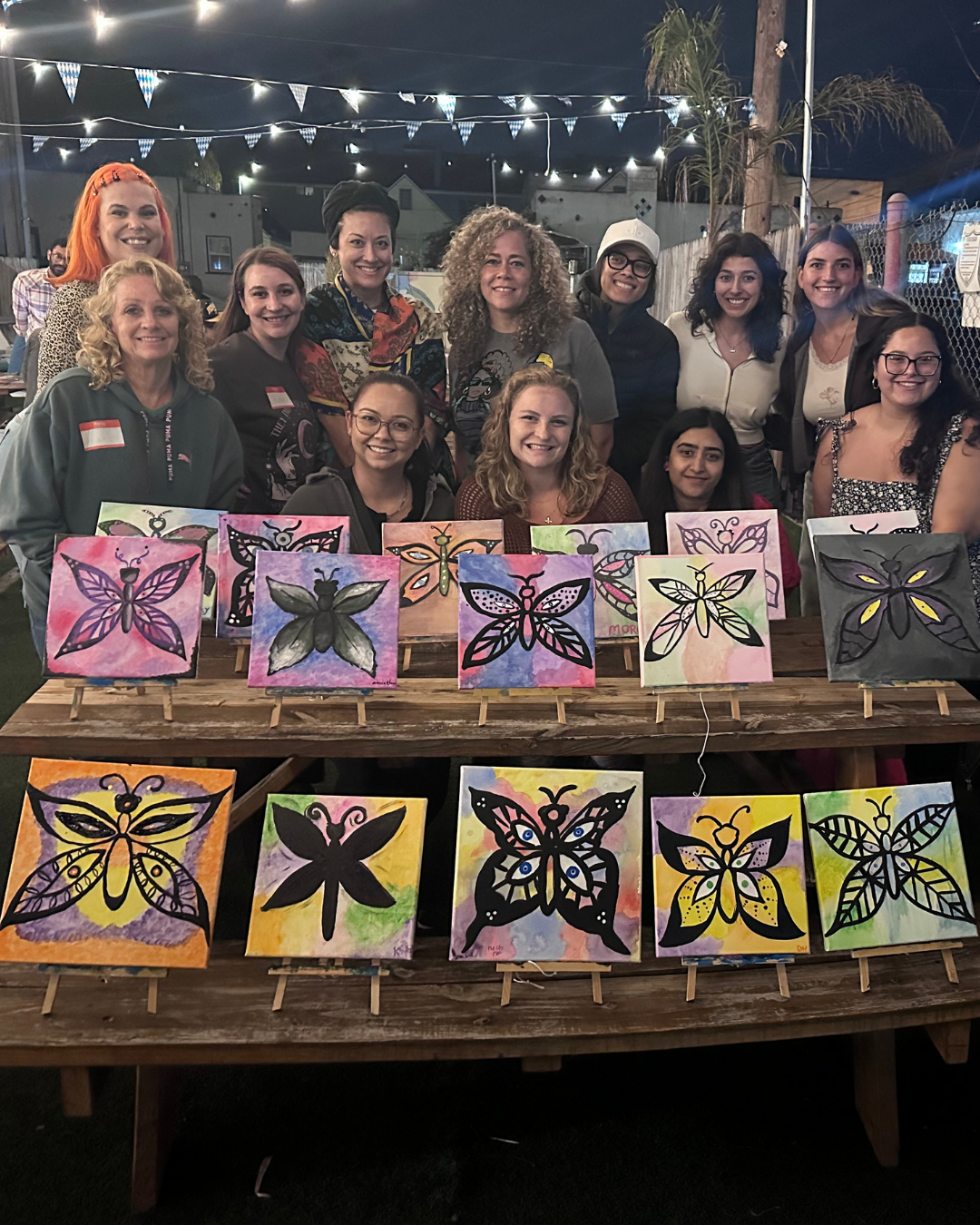 Group of women celebrating at night, displaying butterfly-themed watercolor paintings on small canvases on a wooden table, with string lights and flags overhead.