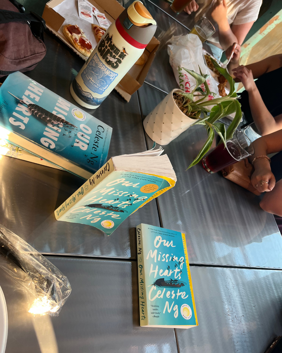 Books titled 'Our Missing Hearts' placed on a table with food and drinks, including pizza, a drink in a plastic cup, and a potted plant.
