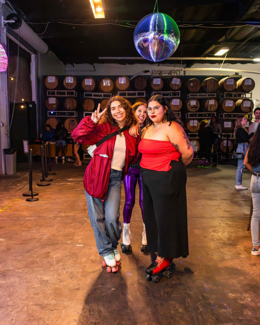Three young women posing with roller skates in a warehouse with barrels and a disco ball overhead, with other people in the background.