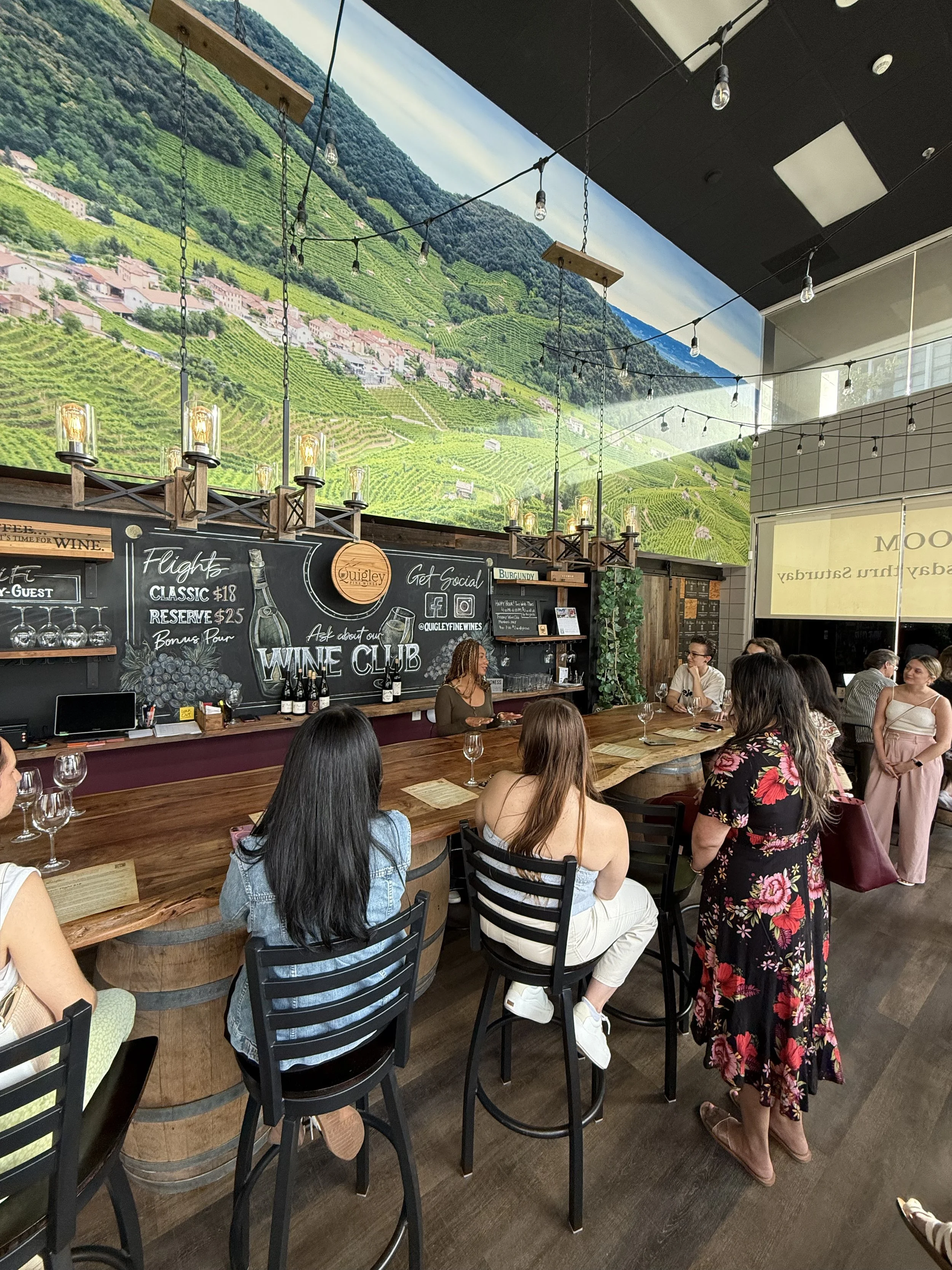 Women gathered at a wine bar for a wine tasting and social event, seated at a wooden counter while a host leads a discussion in a cozy wine club setting with chalkboard menu backdrop.