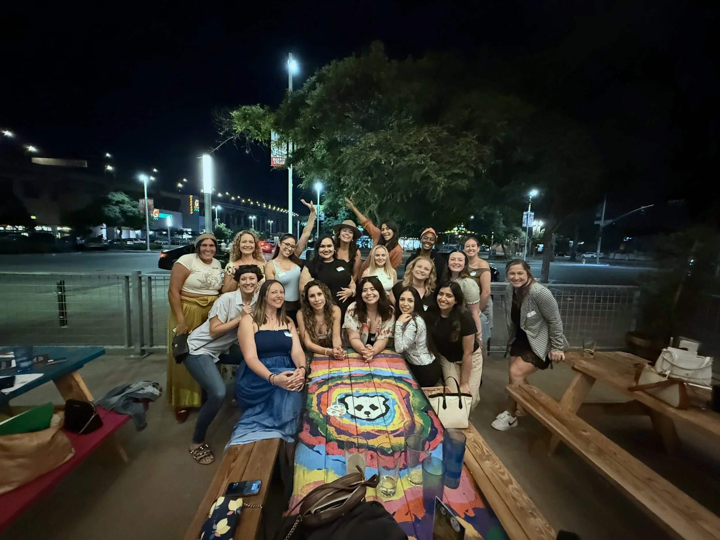 LetsGoGirls San Diego posing outdoors at night in a large group around a colorful picnic table, smiling and celebrating a social gathering.