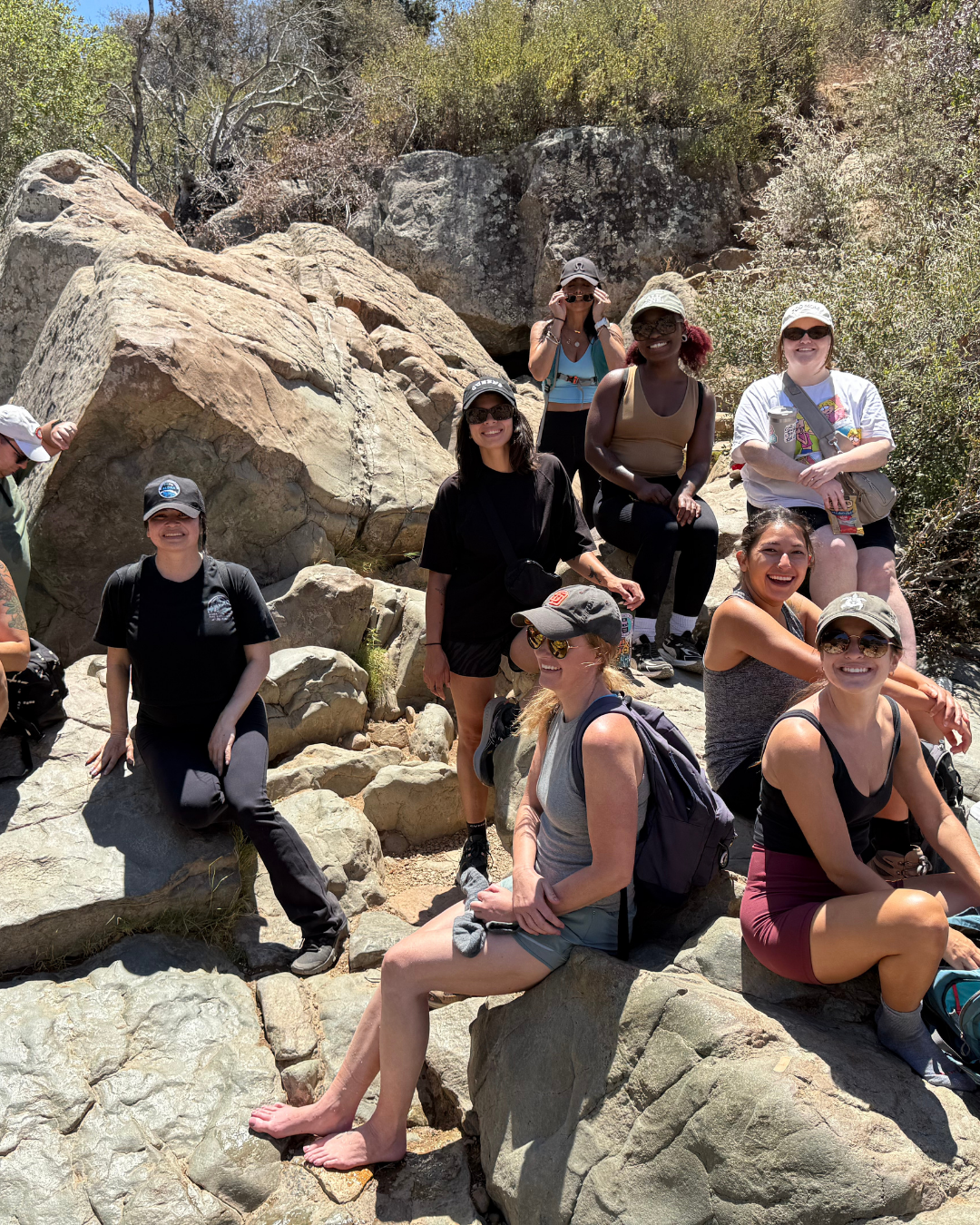 Group of women enjoying a hike on rocky terrain with trees and bushes in the background, some sitting on rocks and others standing, dressed in casual hiking attire and sunglasses.
