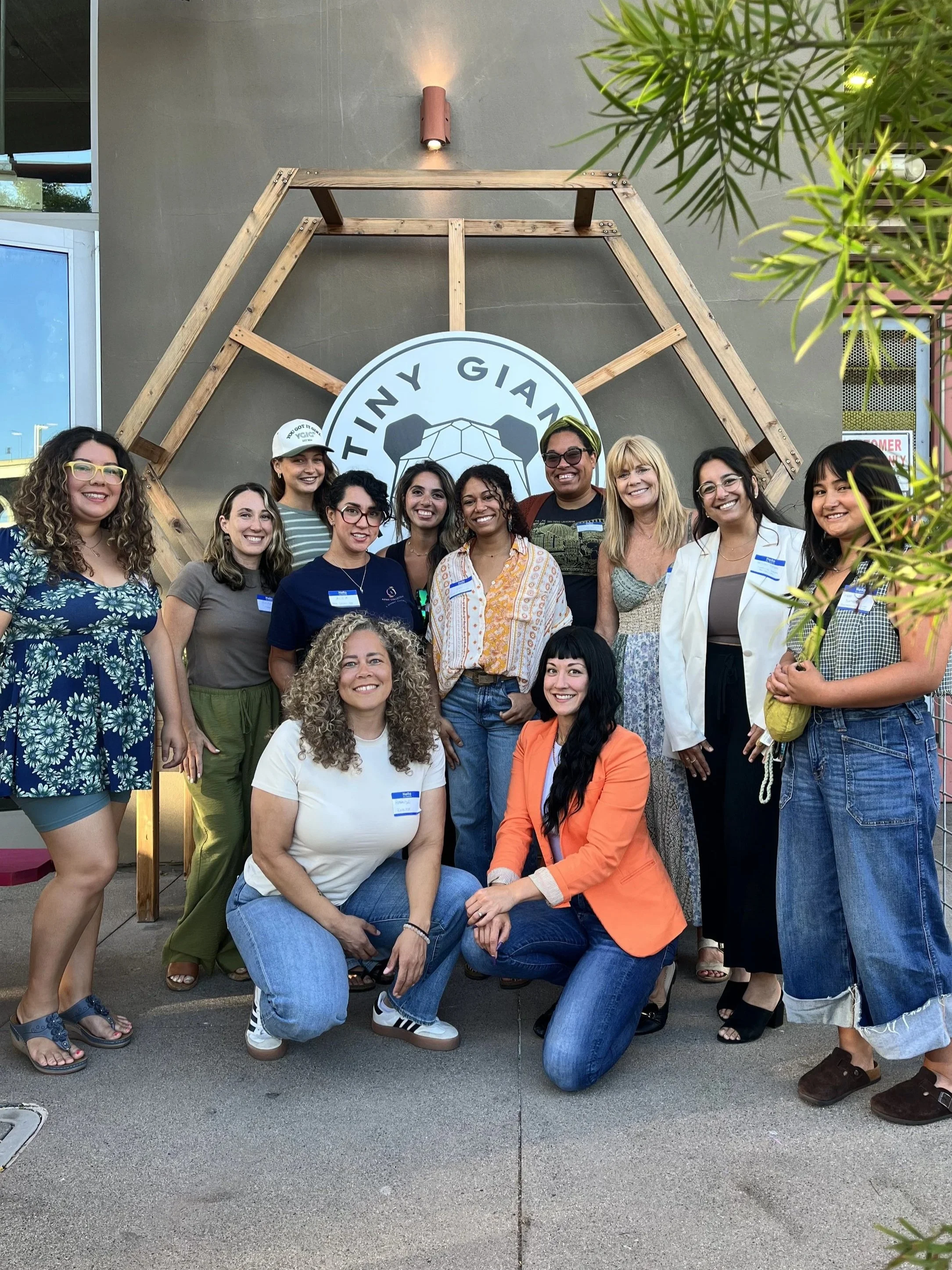 LetsGoGirls San Diego, consisting of diverse women, smiling and posing together in front of the 'Tiny Giants' logo. They are gathered at a community event, radiating empowerment and support for each other.