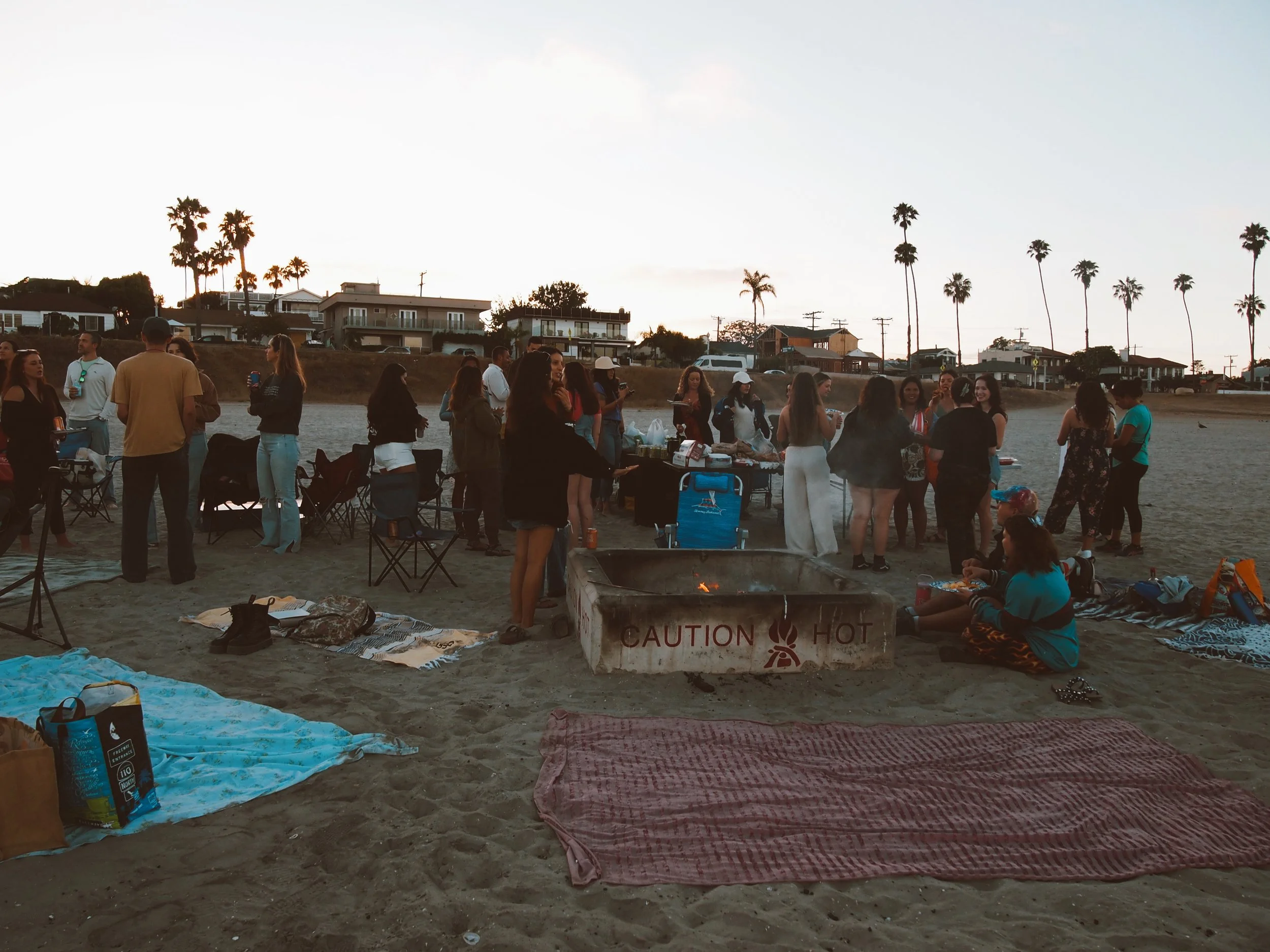 LetsGoGirls San Diego members gathering on the beach at sunset, socializing around a fire pit with blankets and chairs, creating a welcoming atmosphere for friendship, connection, and community.