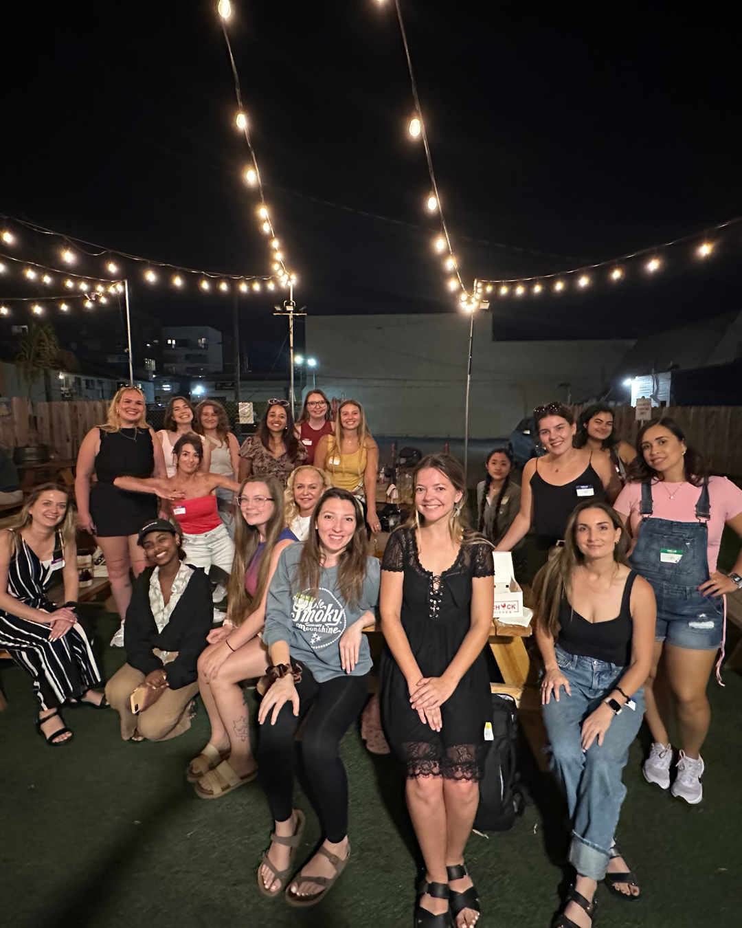 Group of women gathered outdoors at night under string lights, smiling for a photo.