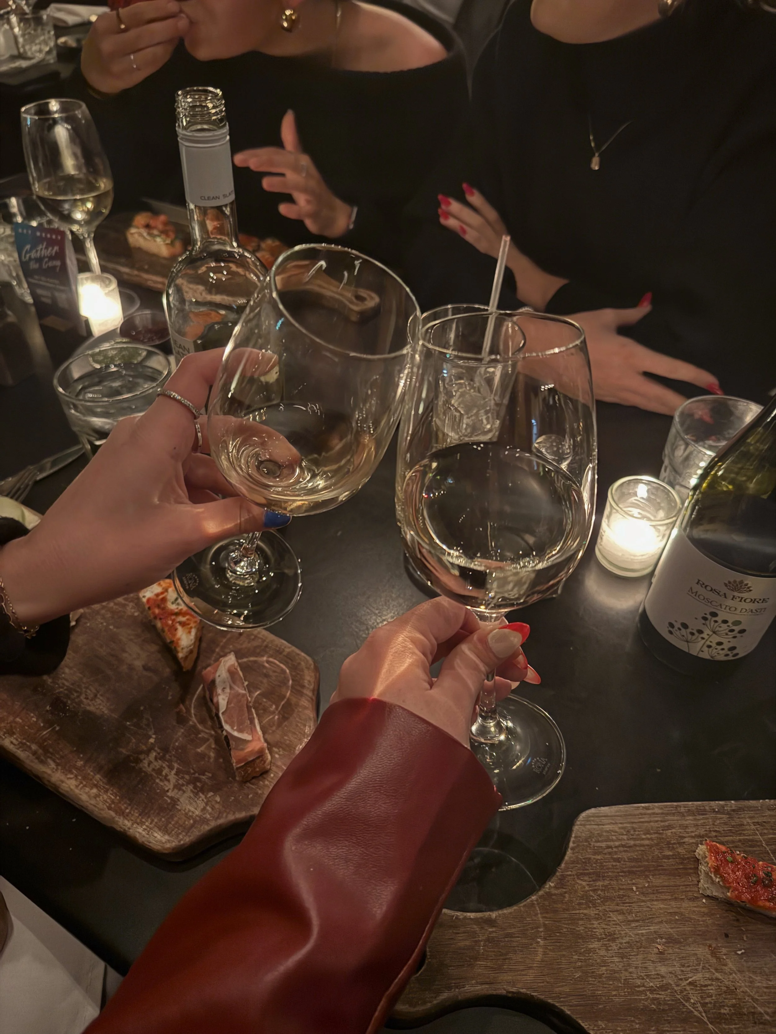 Close-up of two people clinking glasses of wine, with one hand holding a glass and the other holding a clear drink with ice. The background shows soft candlelight and a cozy dining atmosphere with food and drinks on the table.