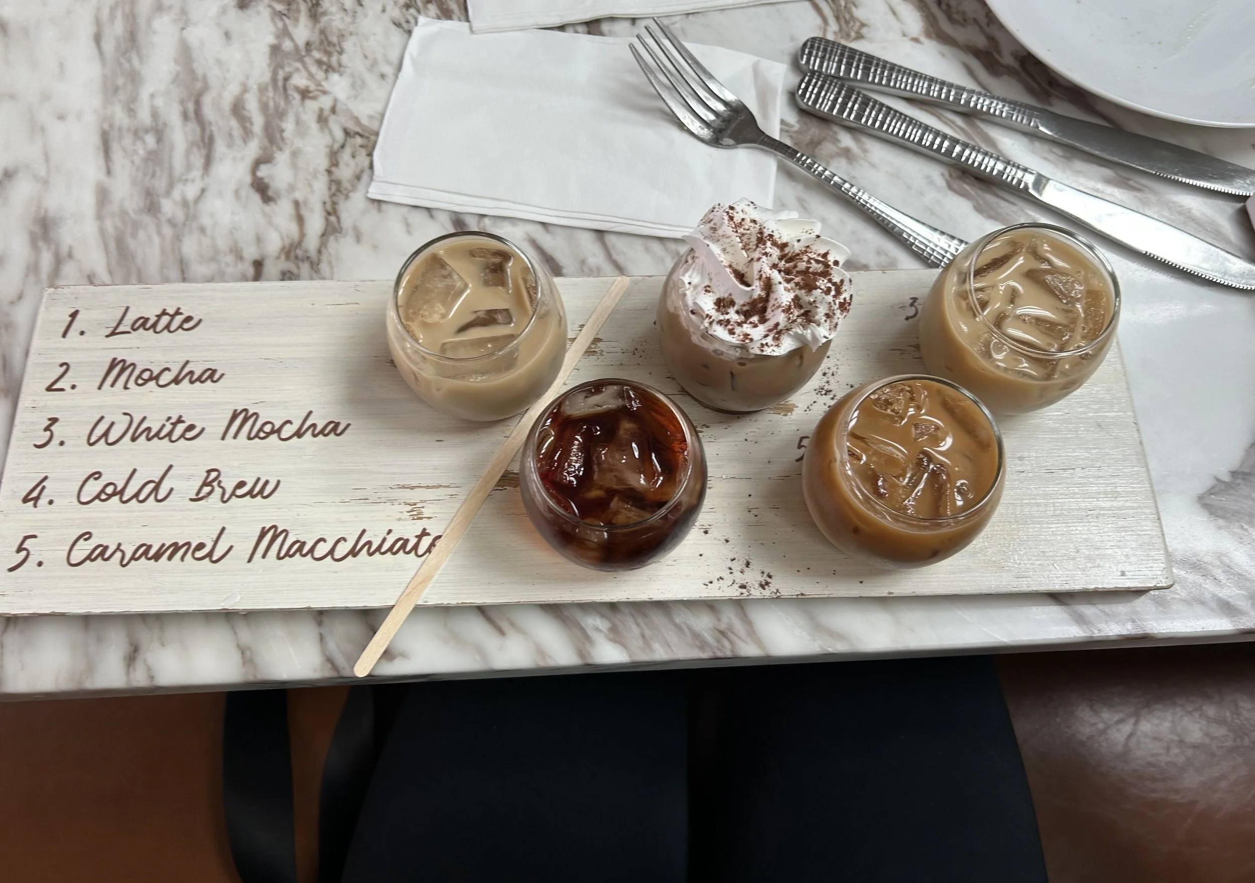 "Coffee tasting experience featuring assorted iced coffee drinks including latte, mocha, white mocha, cold brew, and caramel macchiato arranged on a wooden serving board, showcasing a cozy cafe and social gathering atmosphere.