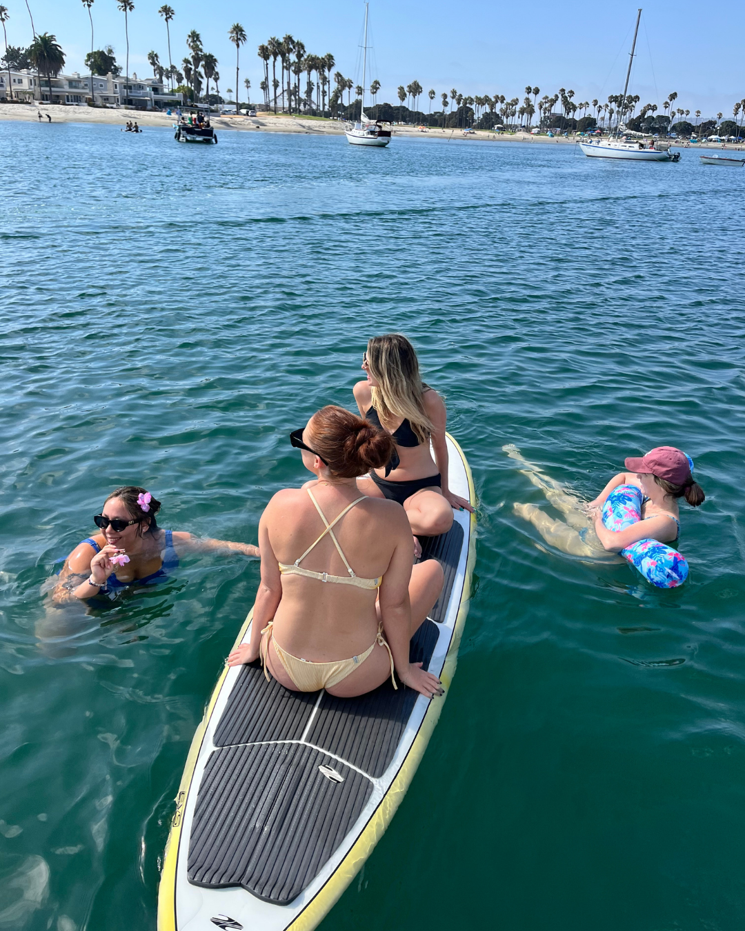 Four women enjoying a day on the water with one sitting on a paddleboard and the other three swimming nearby in a bay with sailboats and palm trees in the background.