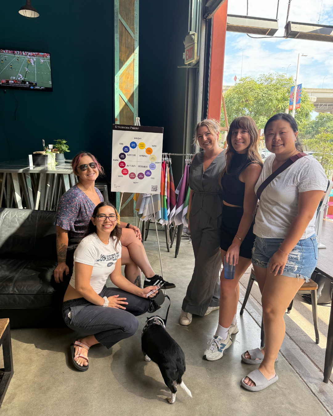 Group of five women with a small black and white dog inside a cafe or store, standing near a color palette chart, with a window showing trees and flags outside.