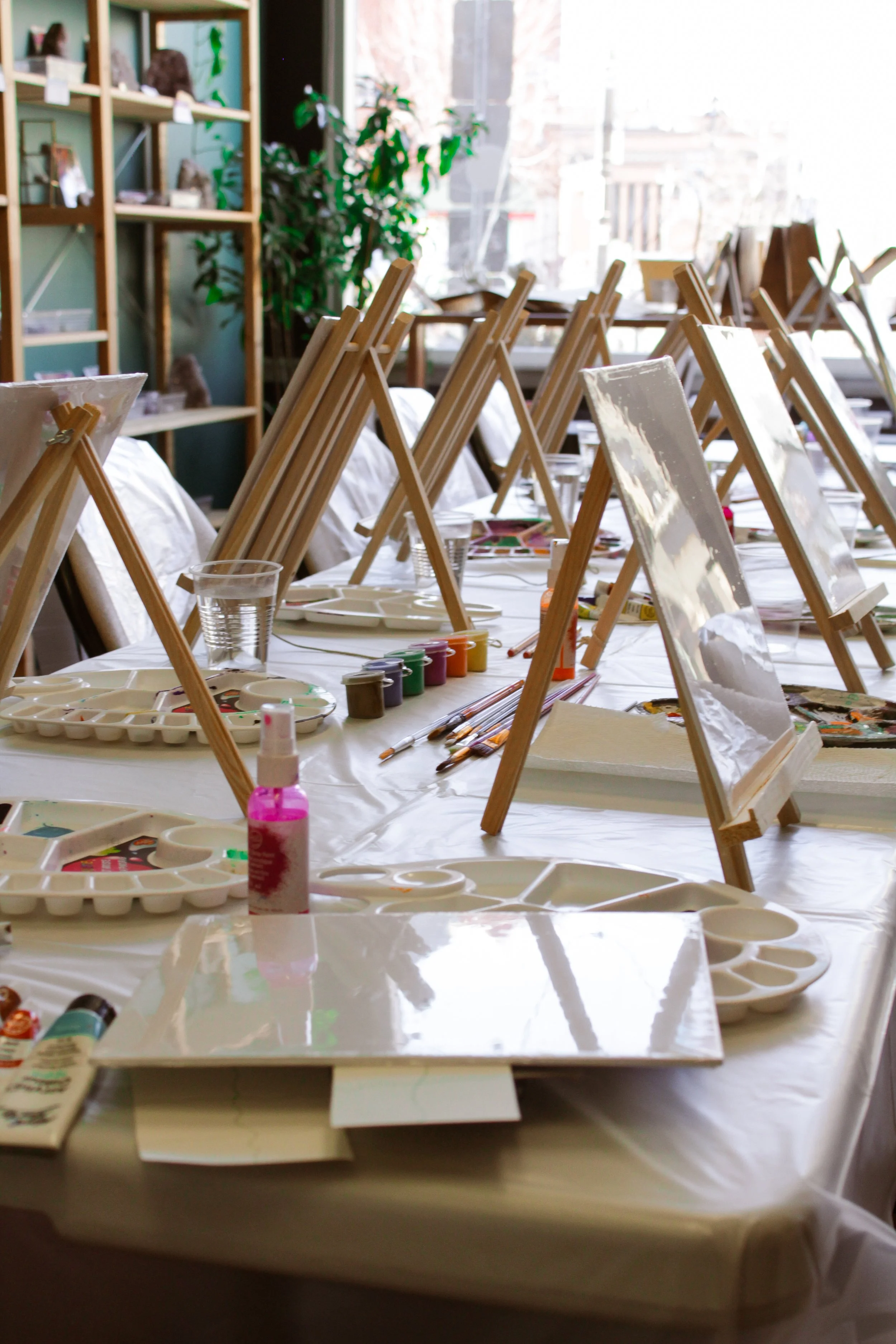 An art studio with tables set up for painting, featuring blank canvases on small easels, paint palettes, brushes, and art supplies, with sunlight coming through large windows.