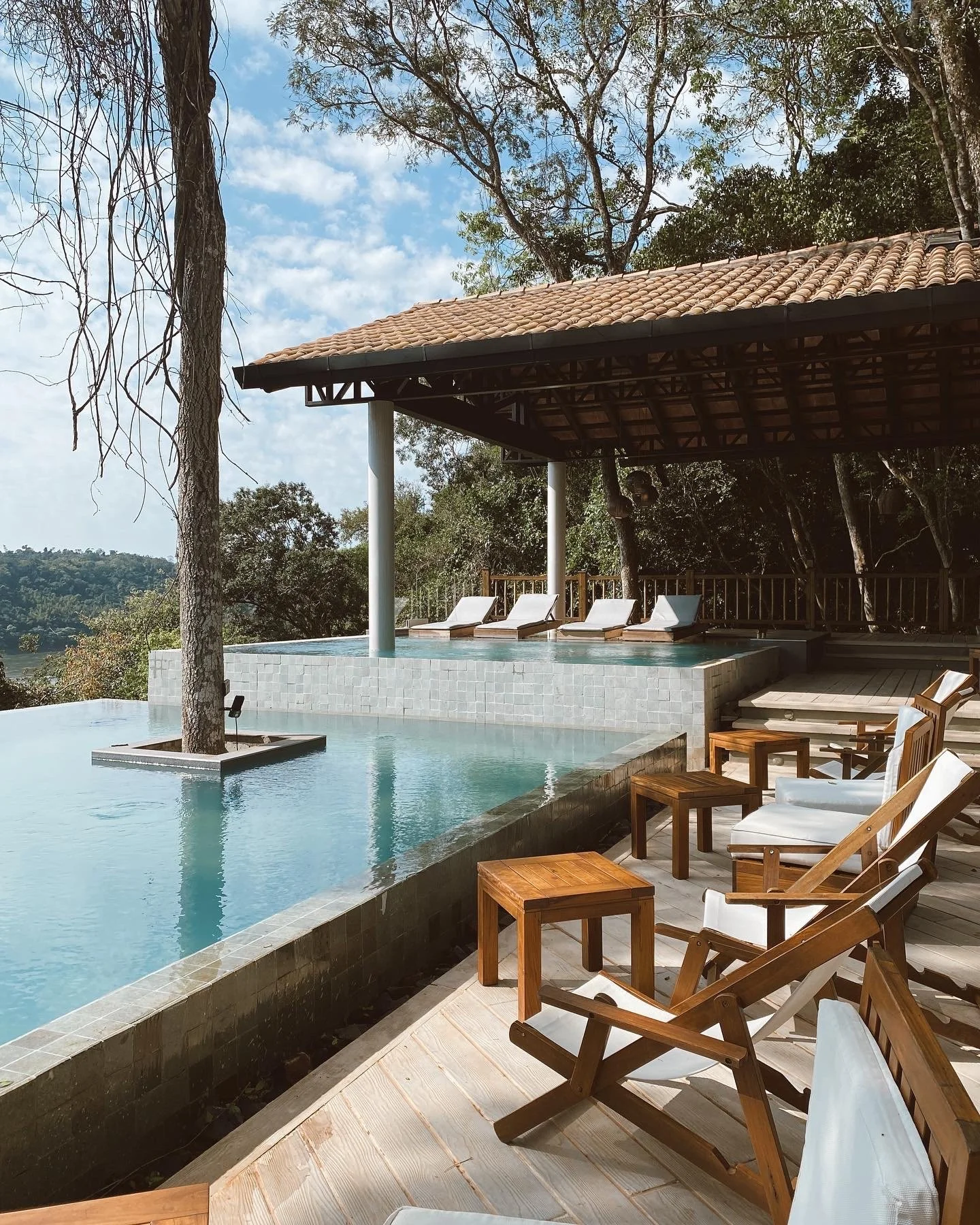 Serene outdoor poolside area with wooden lounge chairs and small tables, overlooking a peaceful landscape. The clear blue water reflects the surrounding greenery and the relaxing, minimalist design of the space.