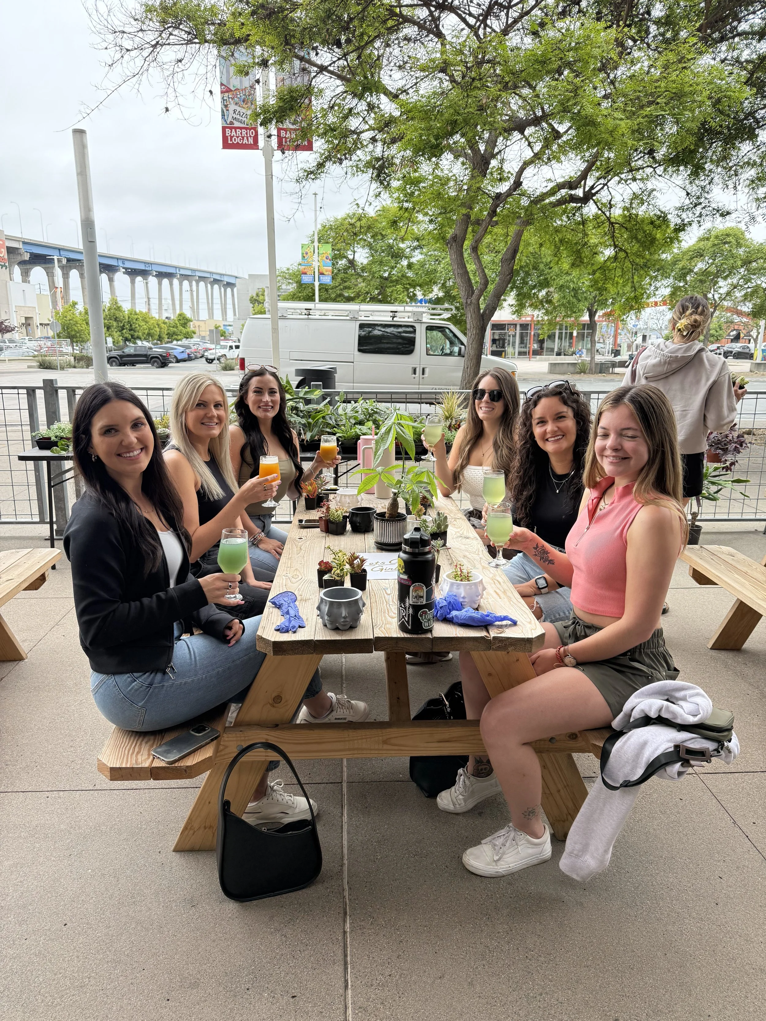 Member of LetsGoGirls San Diego enjoying an outdoor brunch or social gathering, seated at a wooden picnic table with drinks and small plants, smiling at the camera.