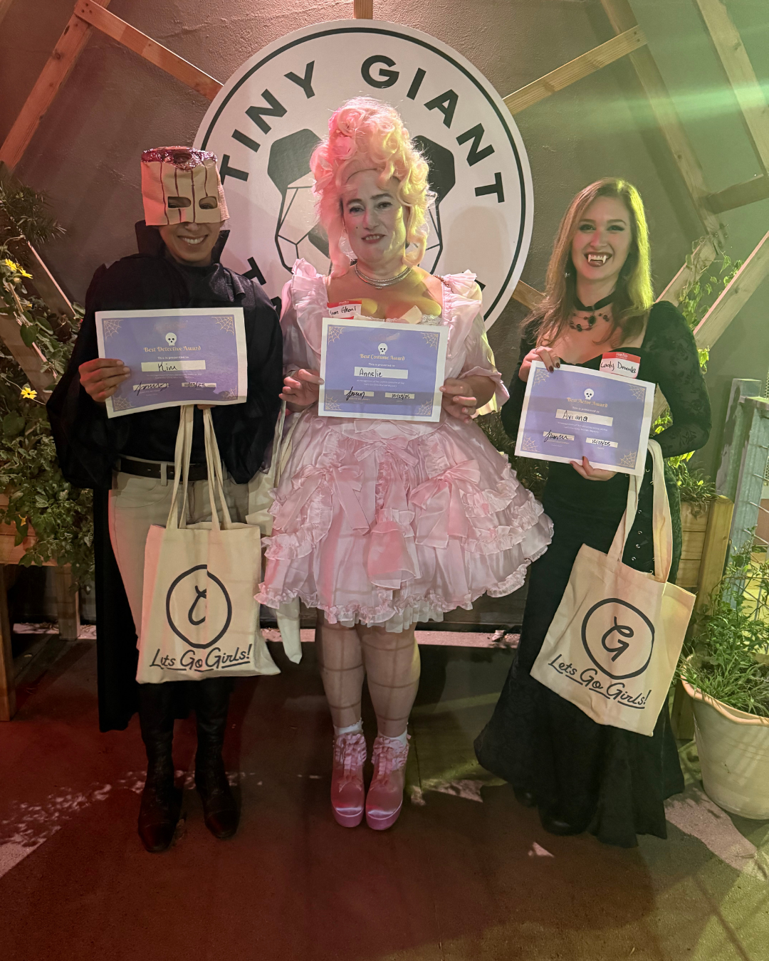 Three women wearing Halloween costumes and holding certificates, standing in front of a backdrop with the words "Tiny Giant". The woman in the center is dressed as a pink princess, and the two women on either side are wearing black Gothic costumes wi