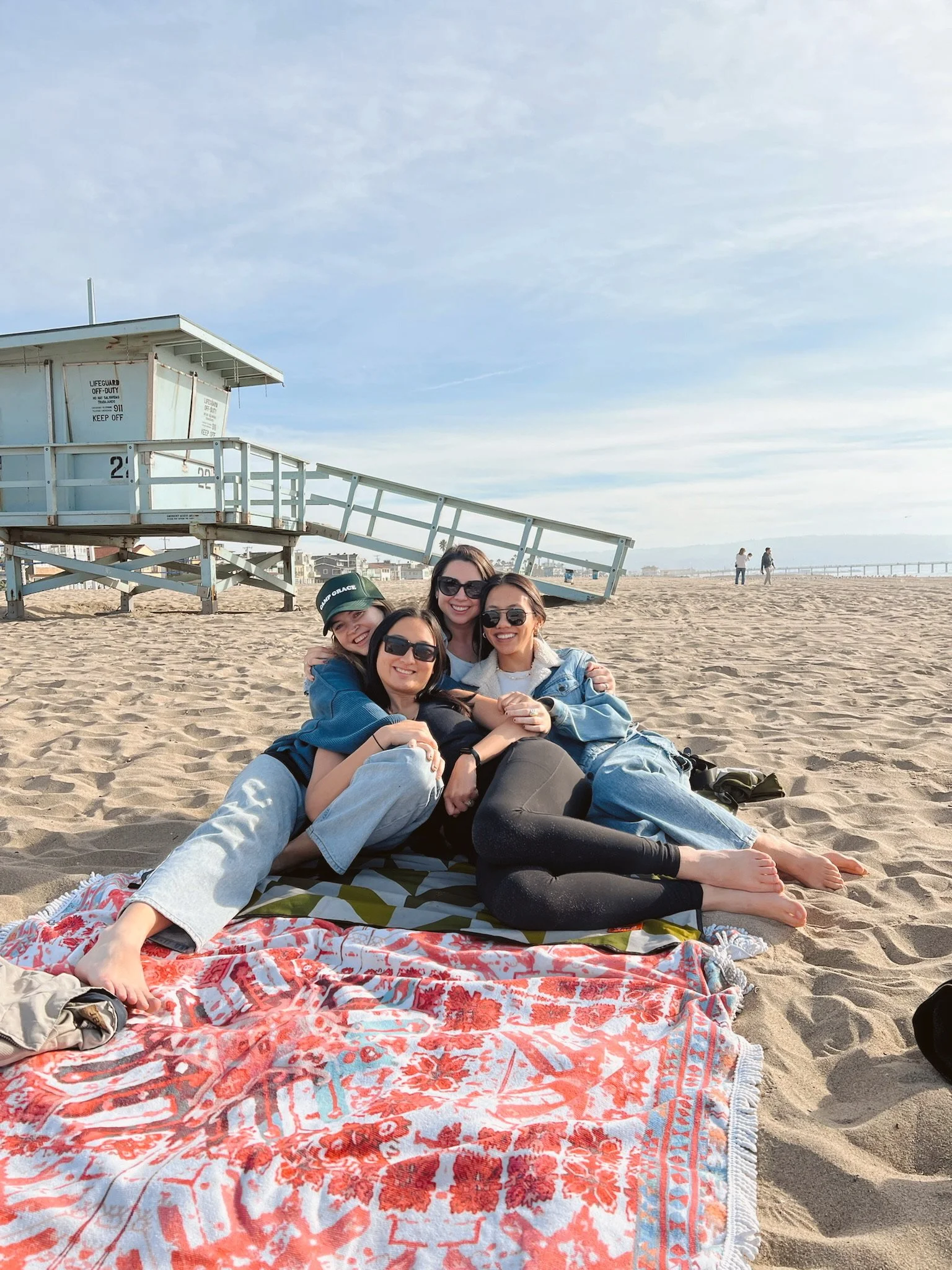 Four women sitting and lying on a colorful beach towel on the sand at the beach, smiling and hugging each other. In the background, there is a lifeguard station and a pier on the ocean.
