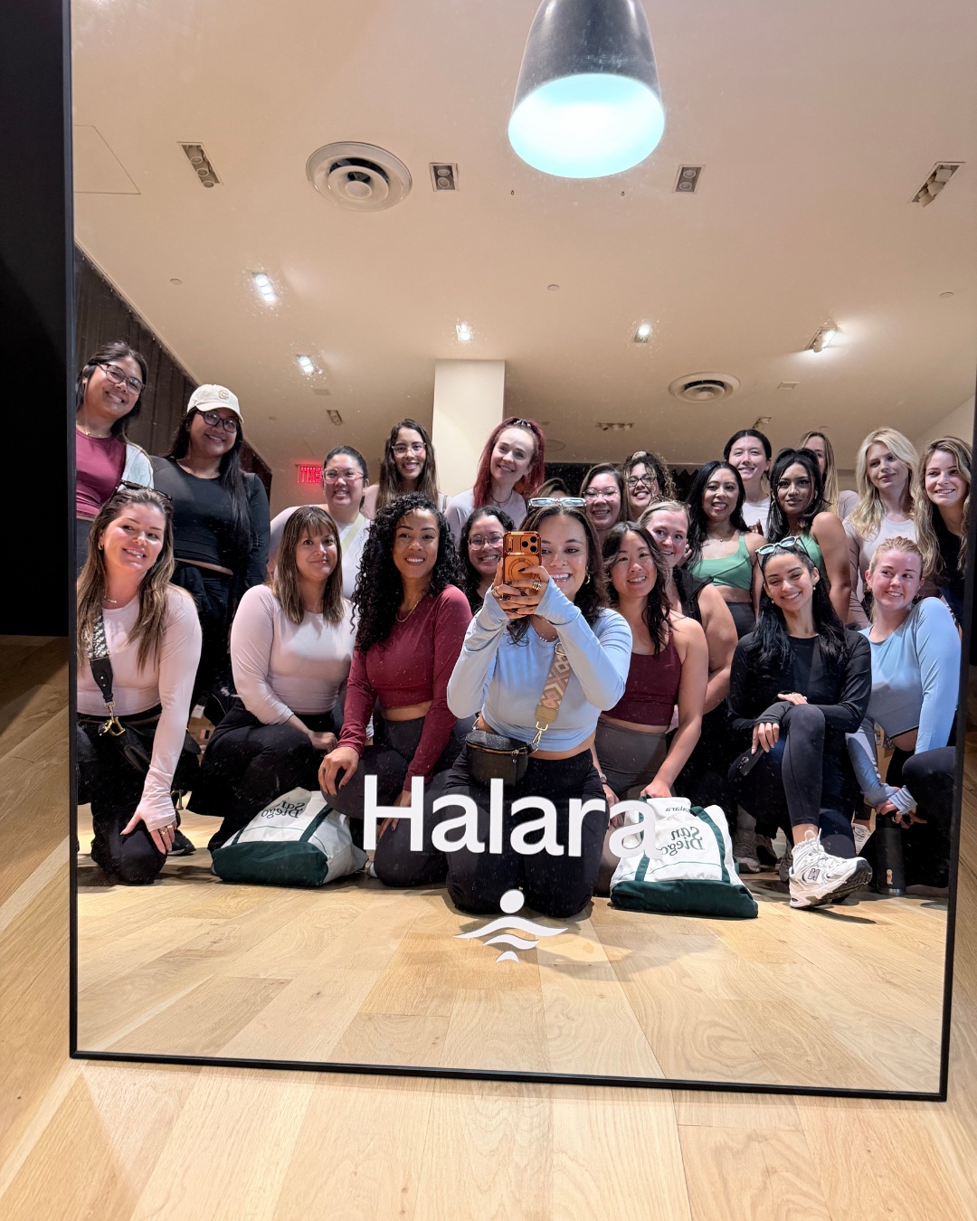 A group of women taking a selfie in front of a mirror with the word 'Halara' on it, in an indoor setting with wooden floors and ceiling lights.