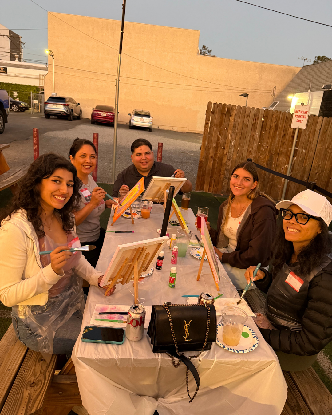 Group of five women sitting at a table outdoors, engaged in painting on canvases, with painting supplies and drinks on the table, in a casual setting during evening.