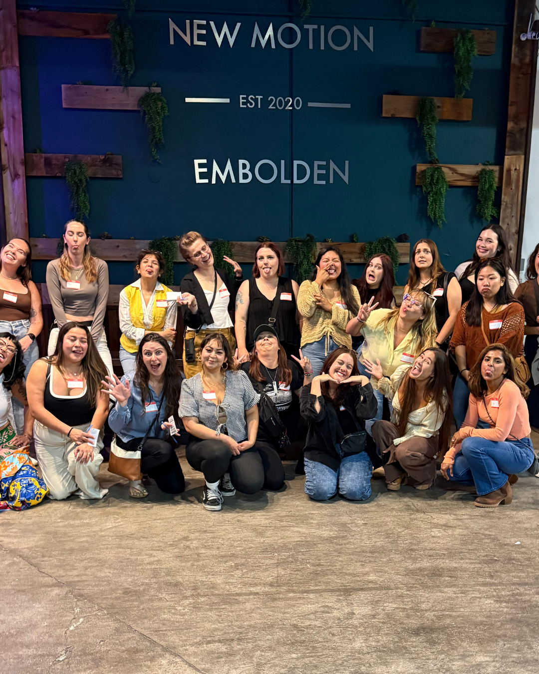 Group of women posing and making funny faces in front of a sign that reads 'New Motion, Est 2020, Embolden' with green foliage and wooden decor in the background.