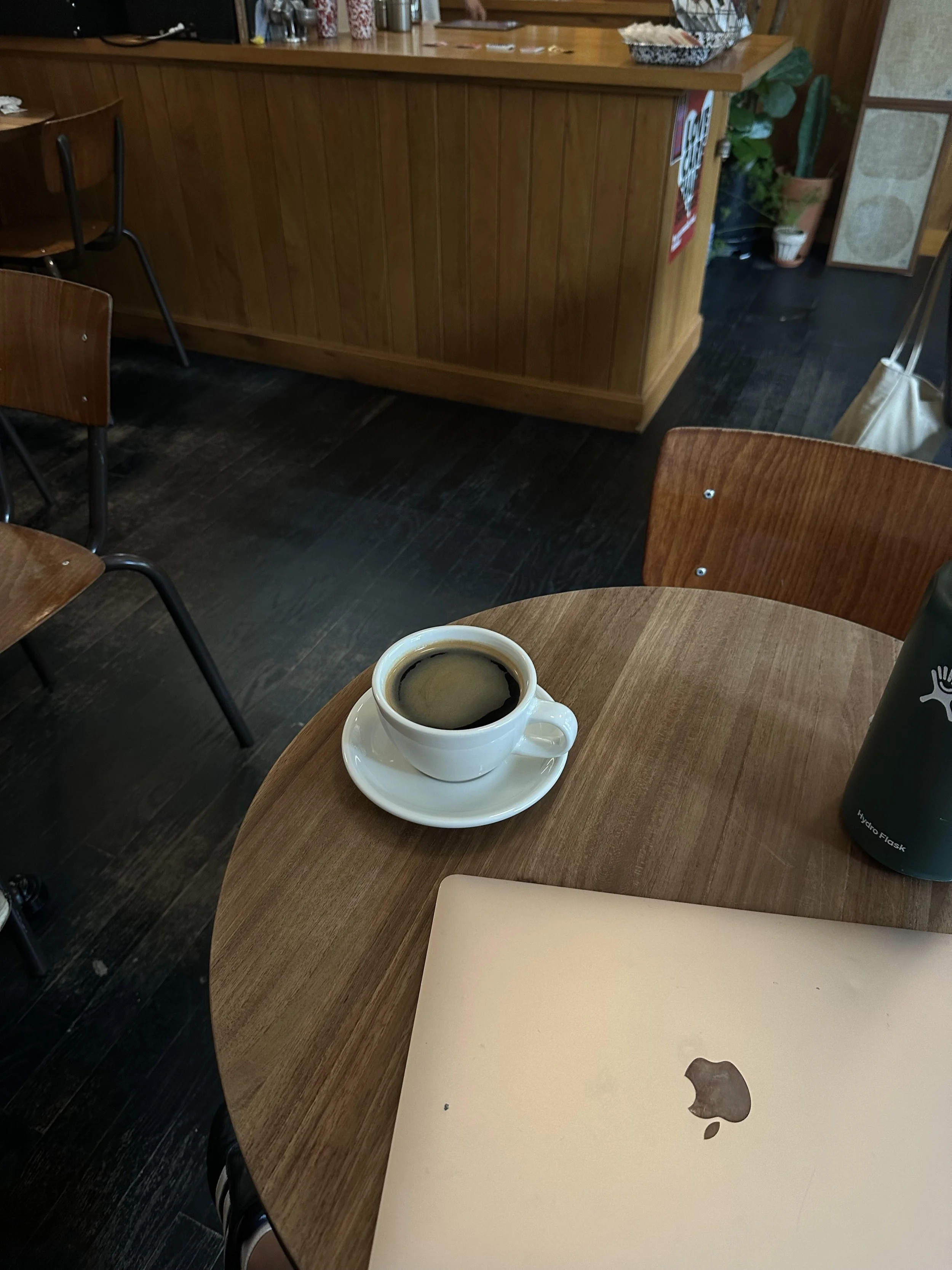 Coffee and laptop workspace on a wooden table, featuring a cup of coffee beside a MacBook in a cozy cafe setting, representing productivity, creativity, and casual community meetups for LetsGoGirls San Diego.
